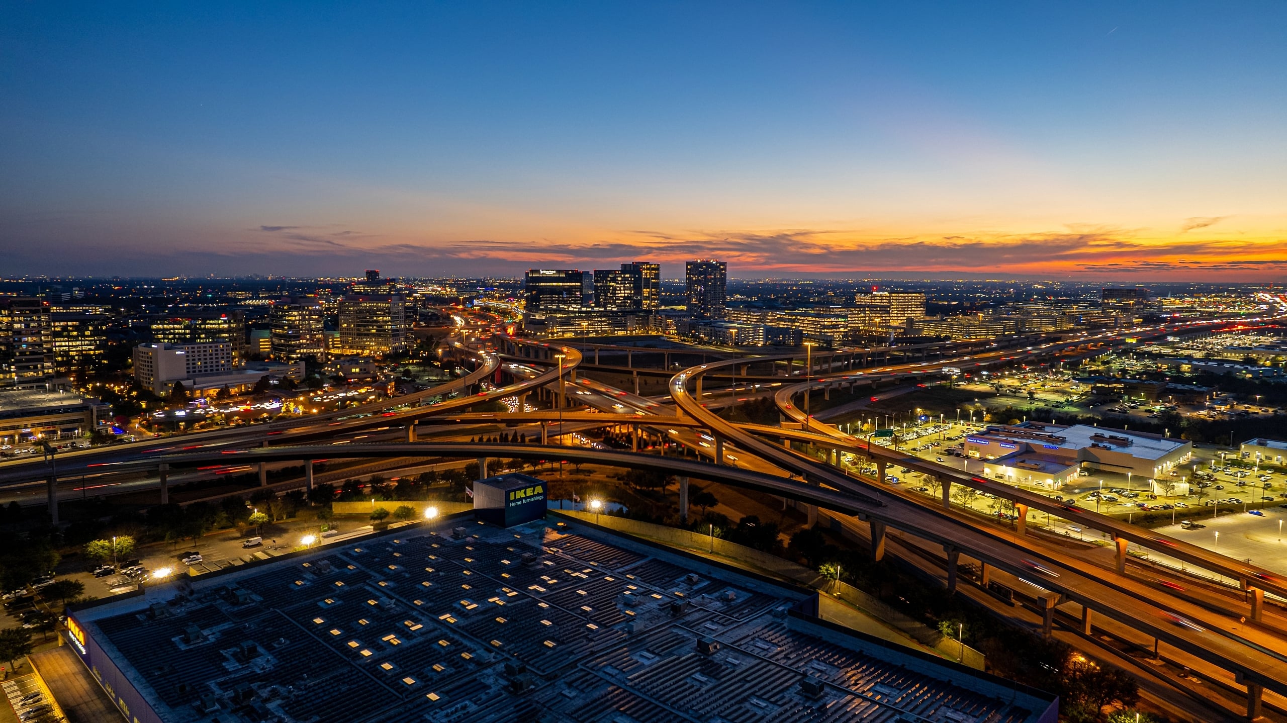 Dramatic nighttime aerial view of a complex highway interchange and a dense urban area with city lights illuminating the scene.