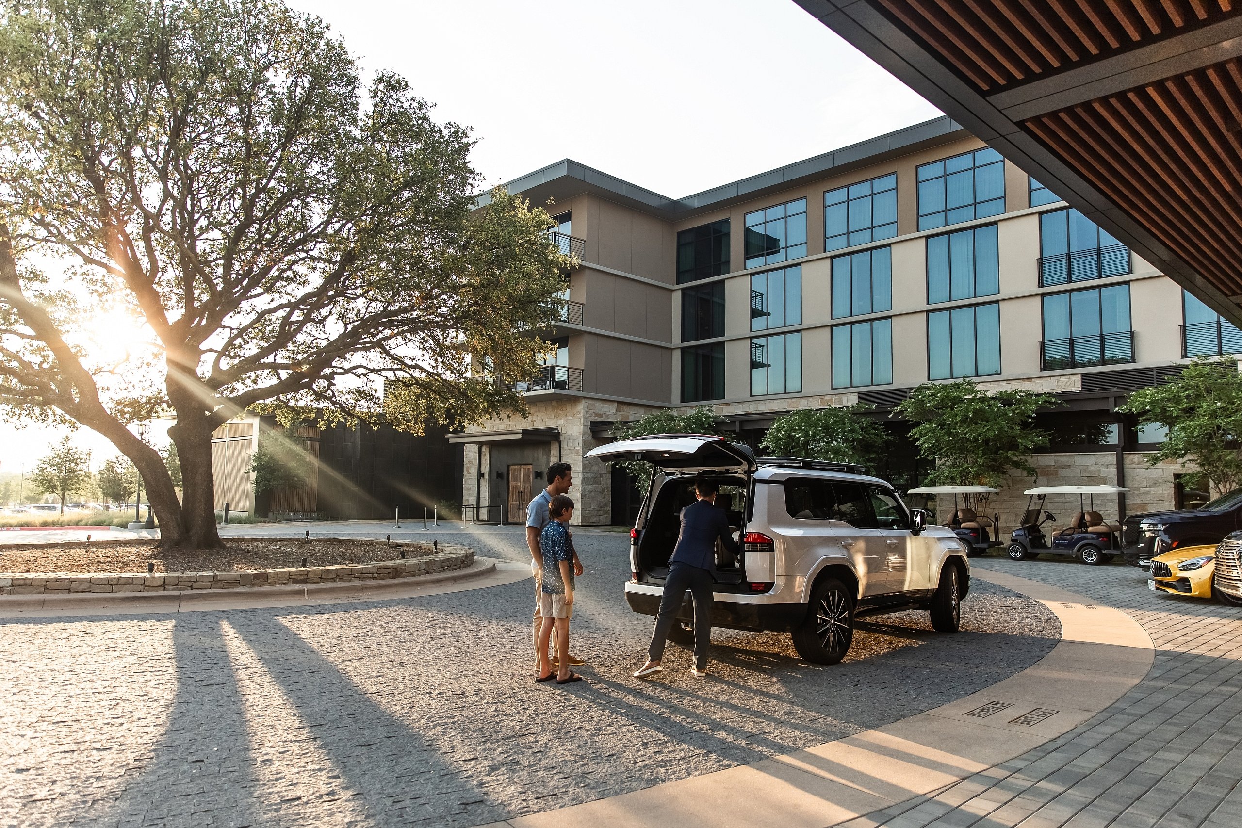 A family loads luggage into a white SUV parked under the hotel's entrance awning at sunset.