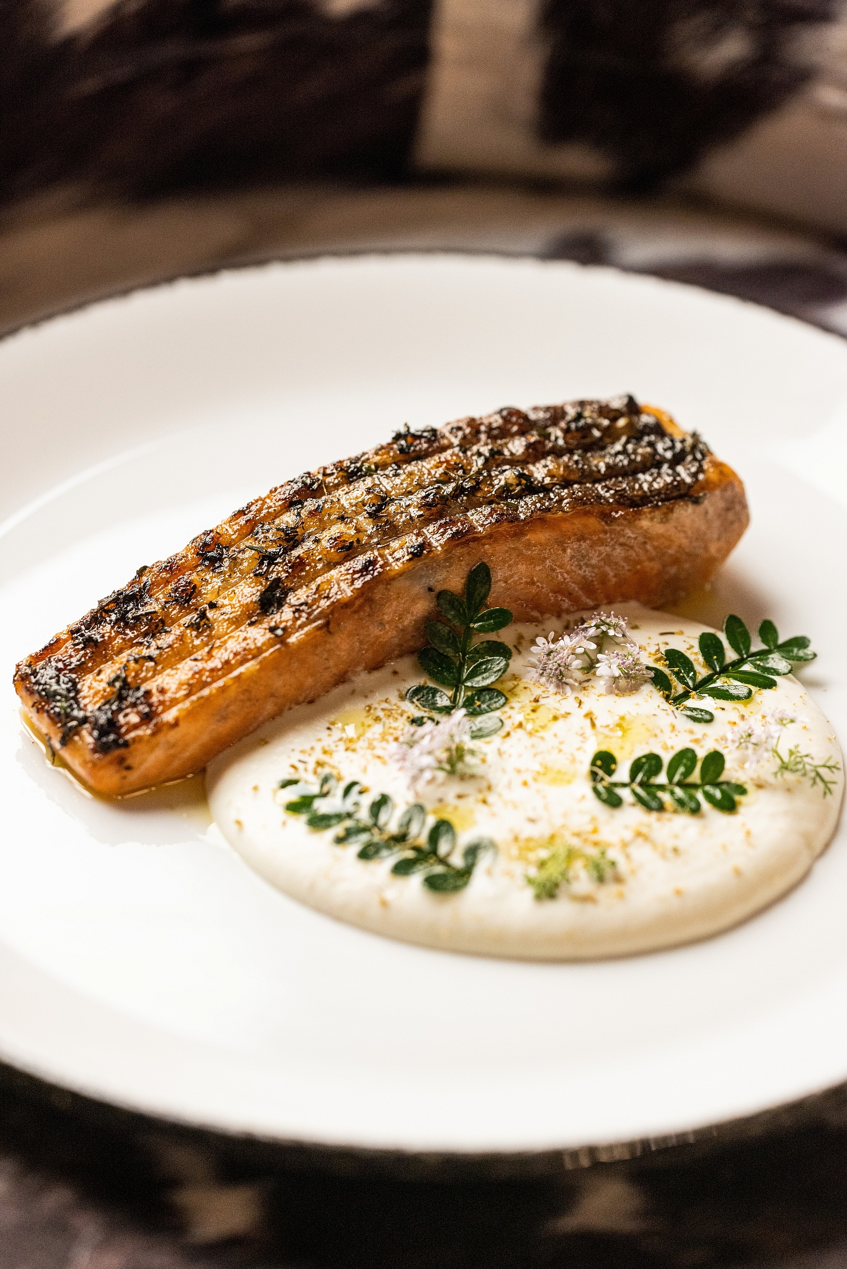 A close-up of a seared salmon fillet served over a creamy white sauce and garnished with delicate herbs and edible flowers.