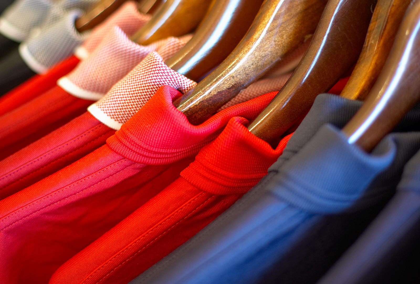 A close-up of brightly colored, collared polo shirts (red, gray, blue) hanging neatly on wooden hangers.