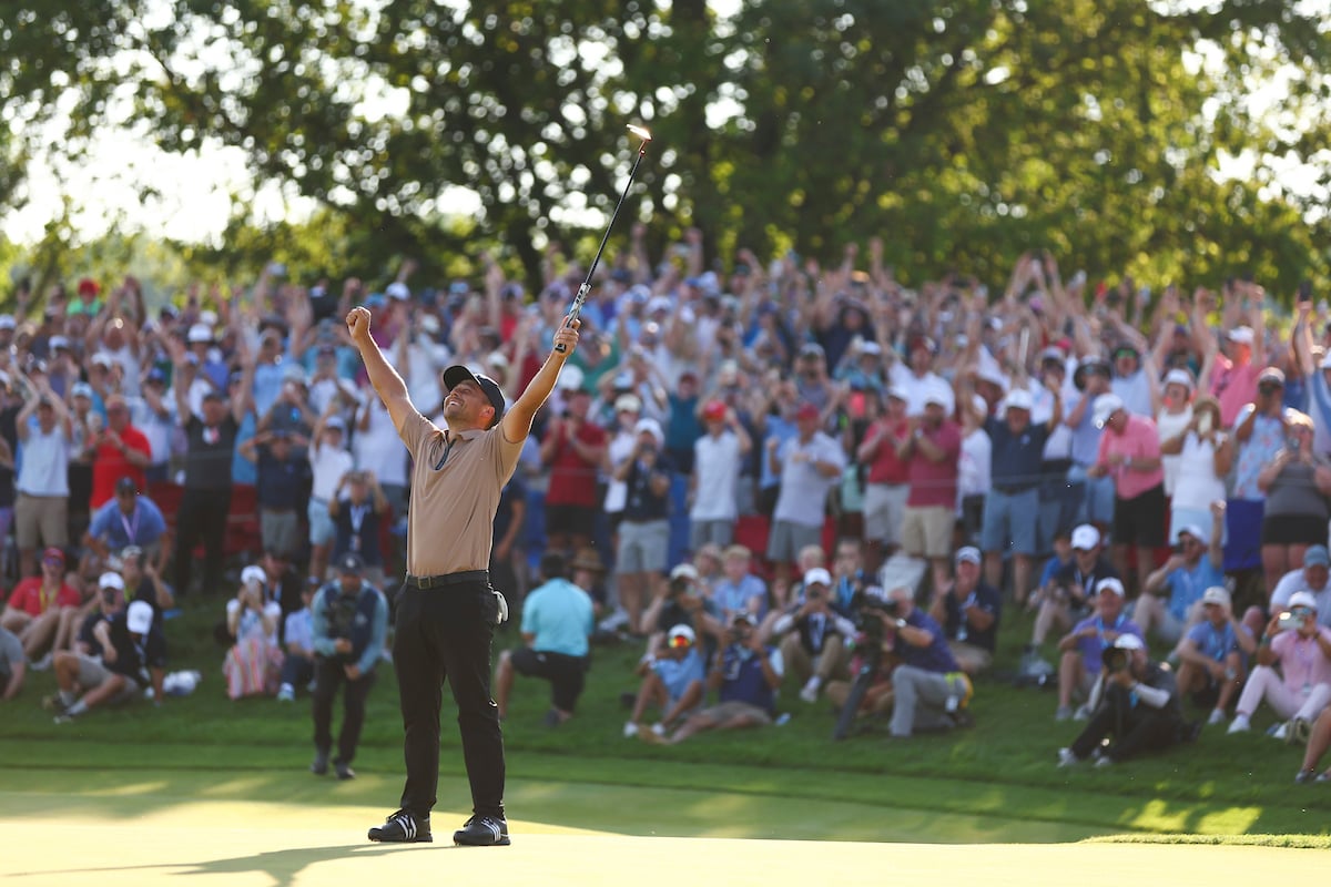 A golfer raises his arms and club in celebration on the green, facing a large, cheering crowd of spectators.