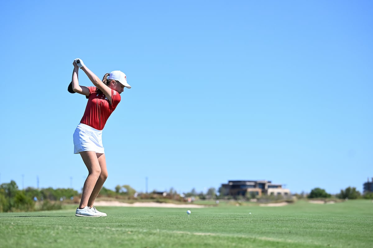 A young female golfer in a red shirt and white skirt completes her swing against a bright, clear blue sky.
