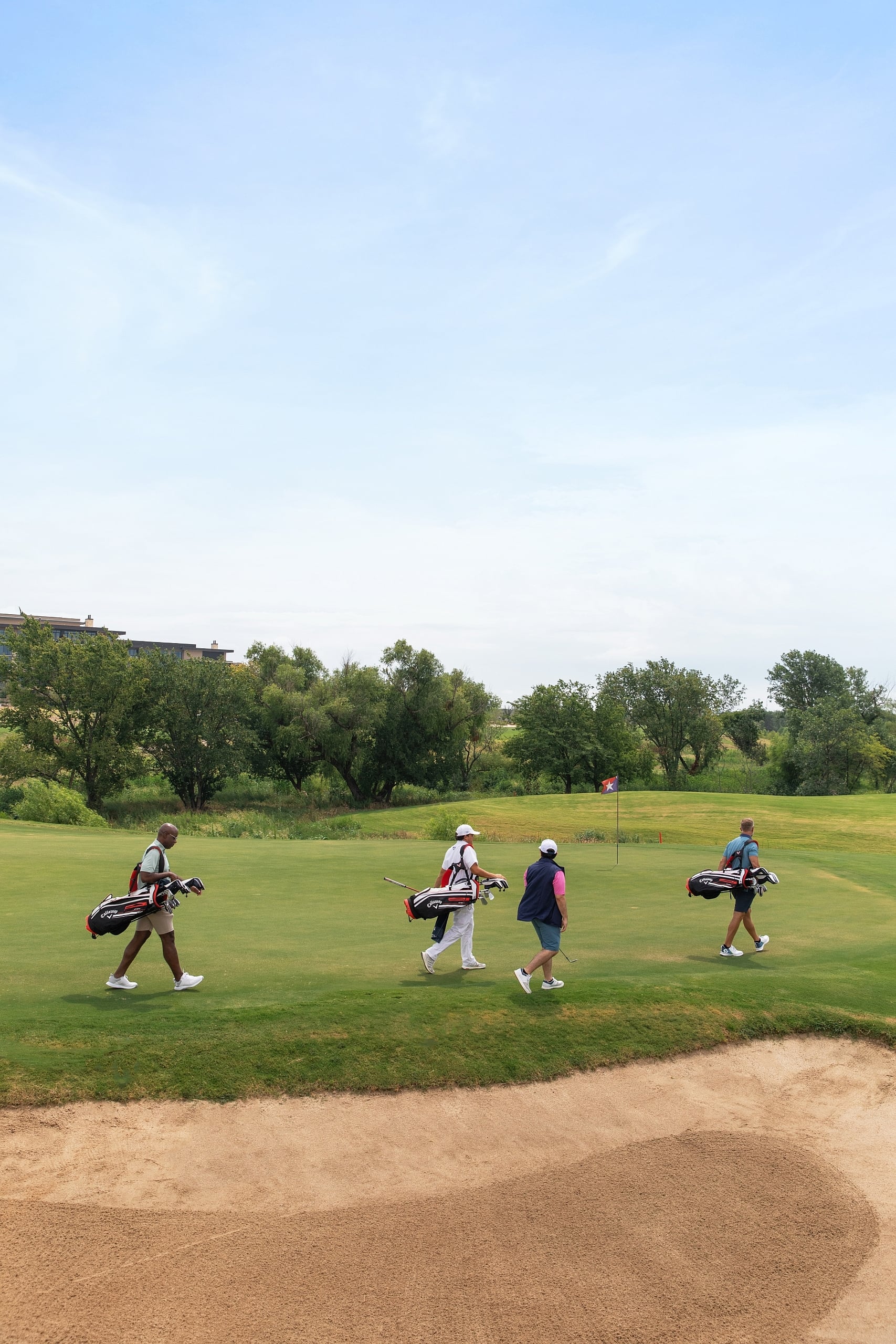 Four golfers and caddies walk across a green fairway past a sand bunker on a sunny day.