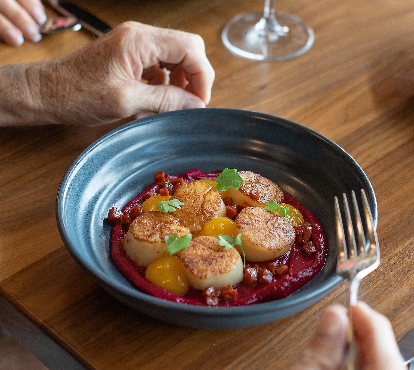 Close-up of pan-seared scallops on a purple puree with yellow garnish, served at a wooden table with white wine.