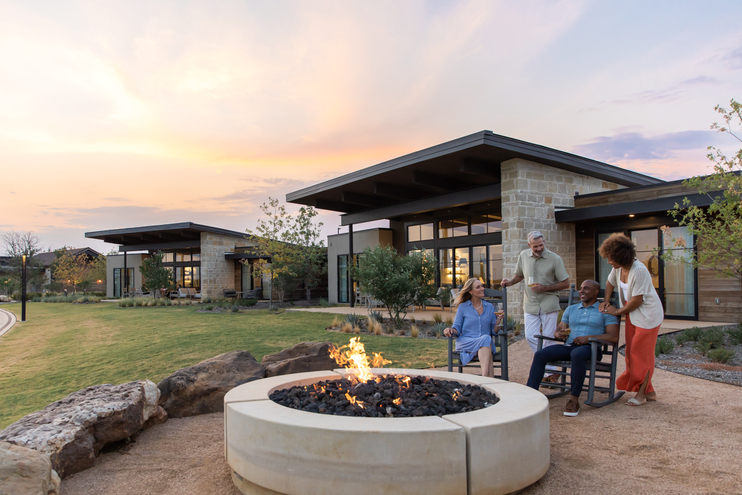 Two couples relax in rocking chairs on a lawn by a stone fire pit, with modern stone and wood resort buildings behind them.