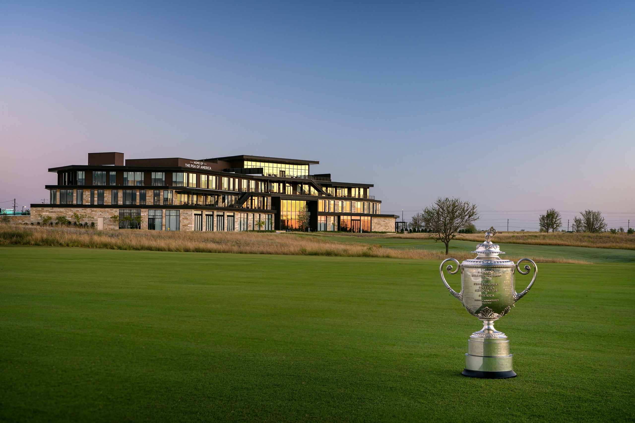 A silver golf trophy stands on the green fairway, with the modern PGA Frisco headquarters building in the background at sunrise.
