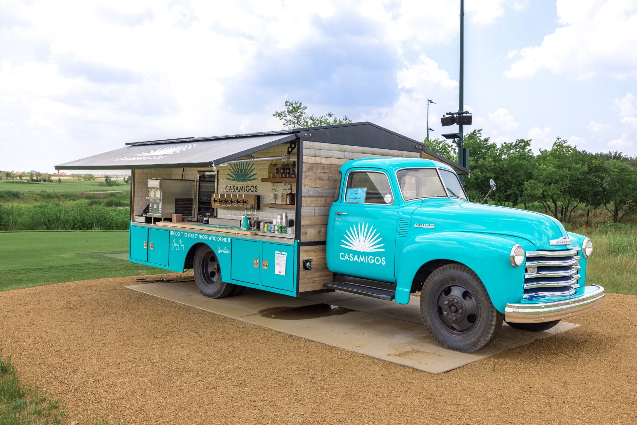 A vintage turquoise Chevrolet pickup truck converted into a Casamigos tequila mobile bar, parked near the golf course.