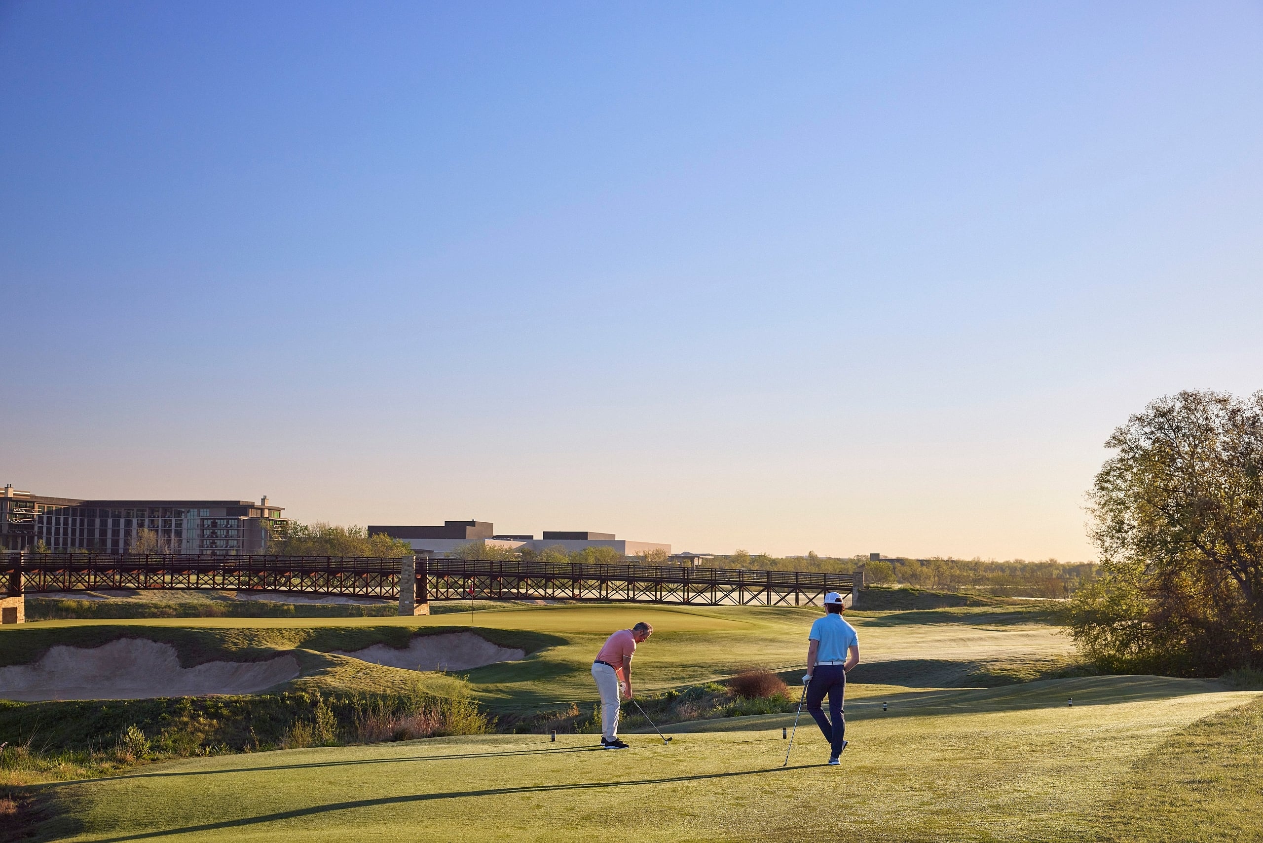 Two golfers putt on a green at sunrise, with the hotel complex and a long bridge visible in the hazy distance.