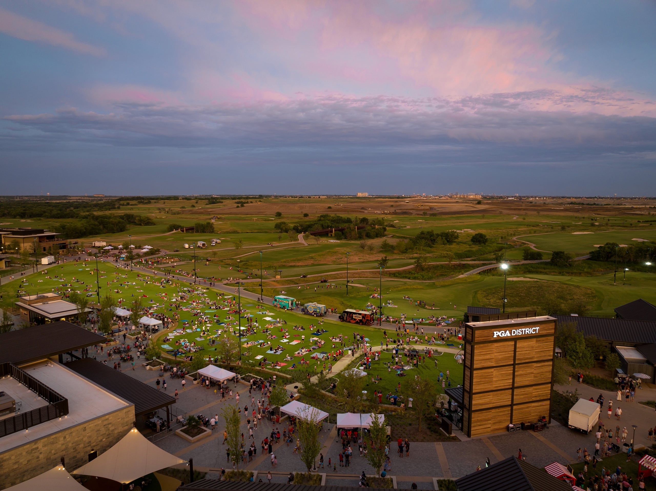 Elevated view of a large outdoor gathering on a vast lawn (PGA District), surrounded by resort buildings at dusk.