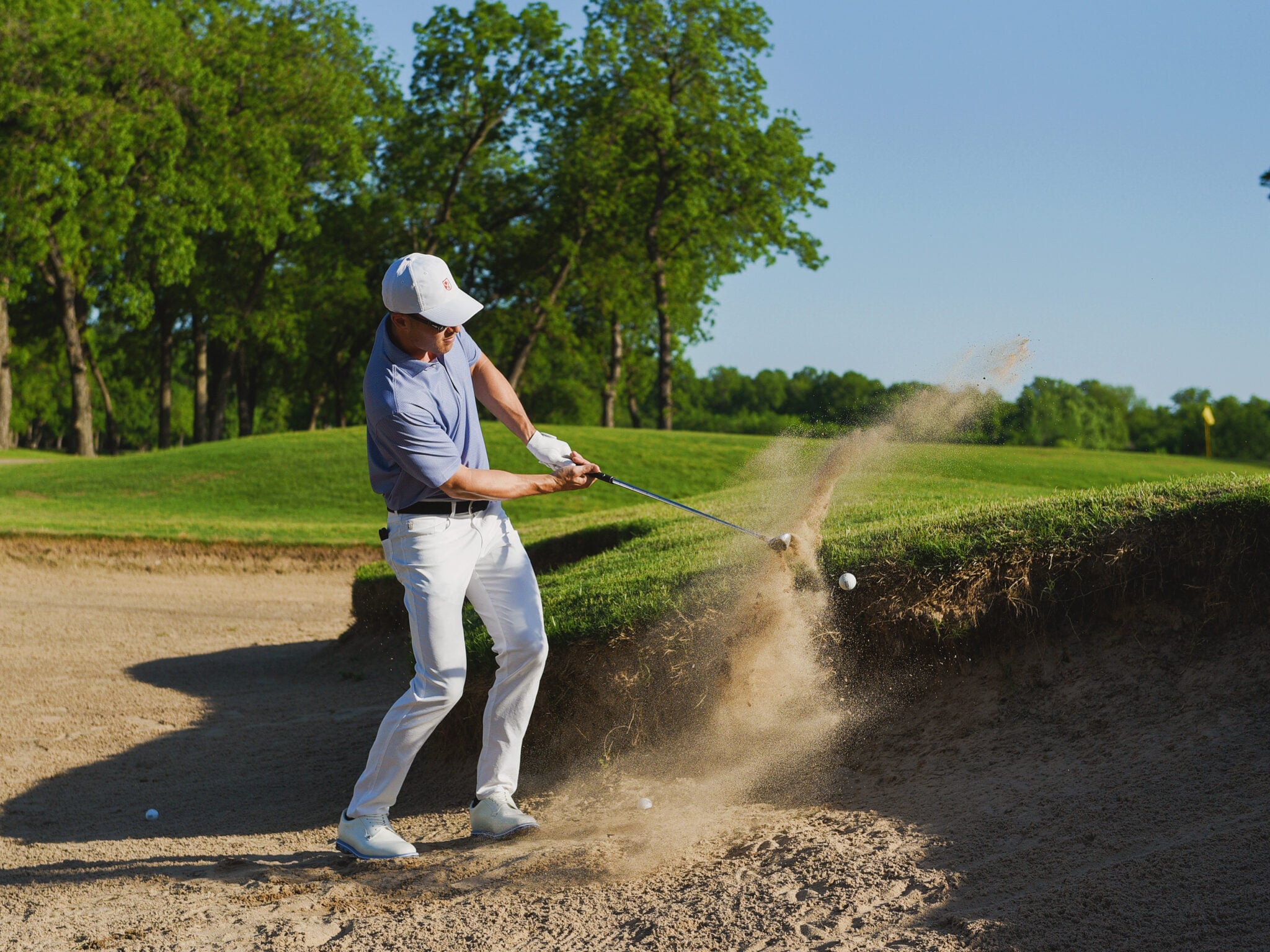 A golfer hits a dynamic bunker shot, sending a large plume of sand and dust flying on the golf course.