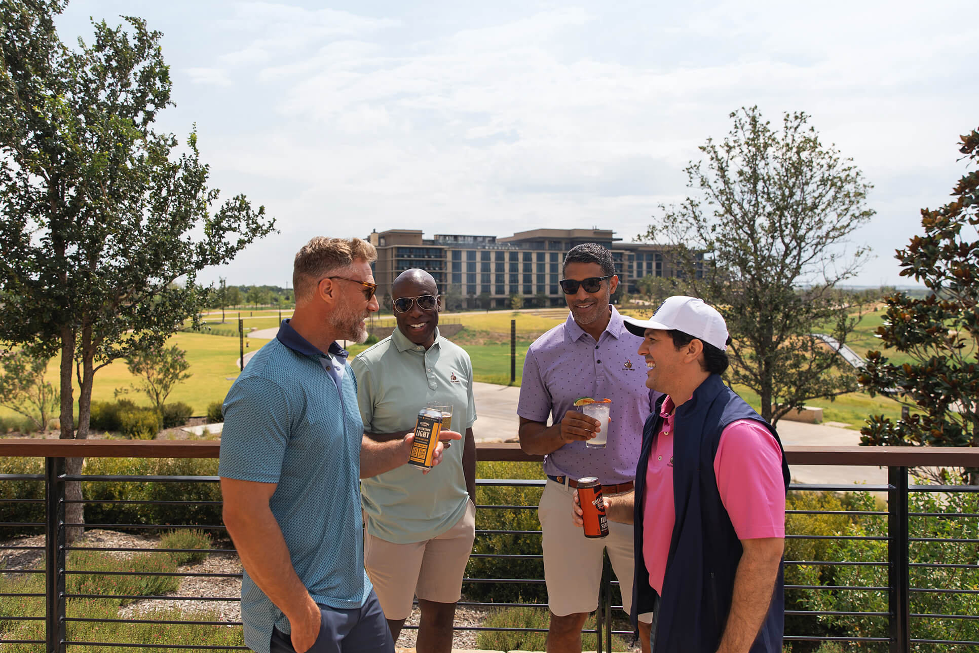 A group of four men laughing and enjoying canned drinks and cocktails on a sunny balcony overlooking the resort grounds.