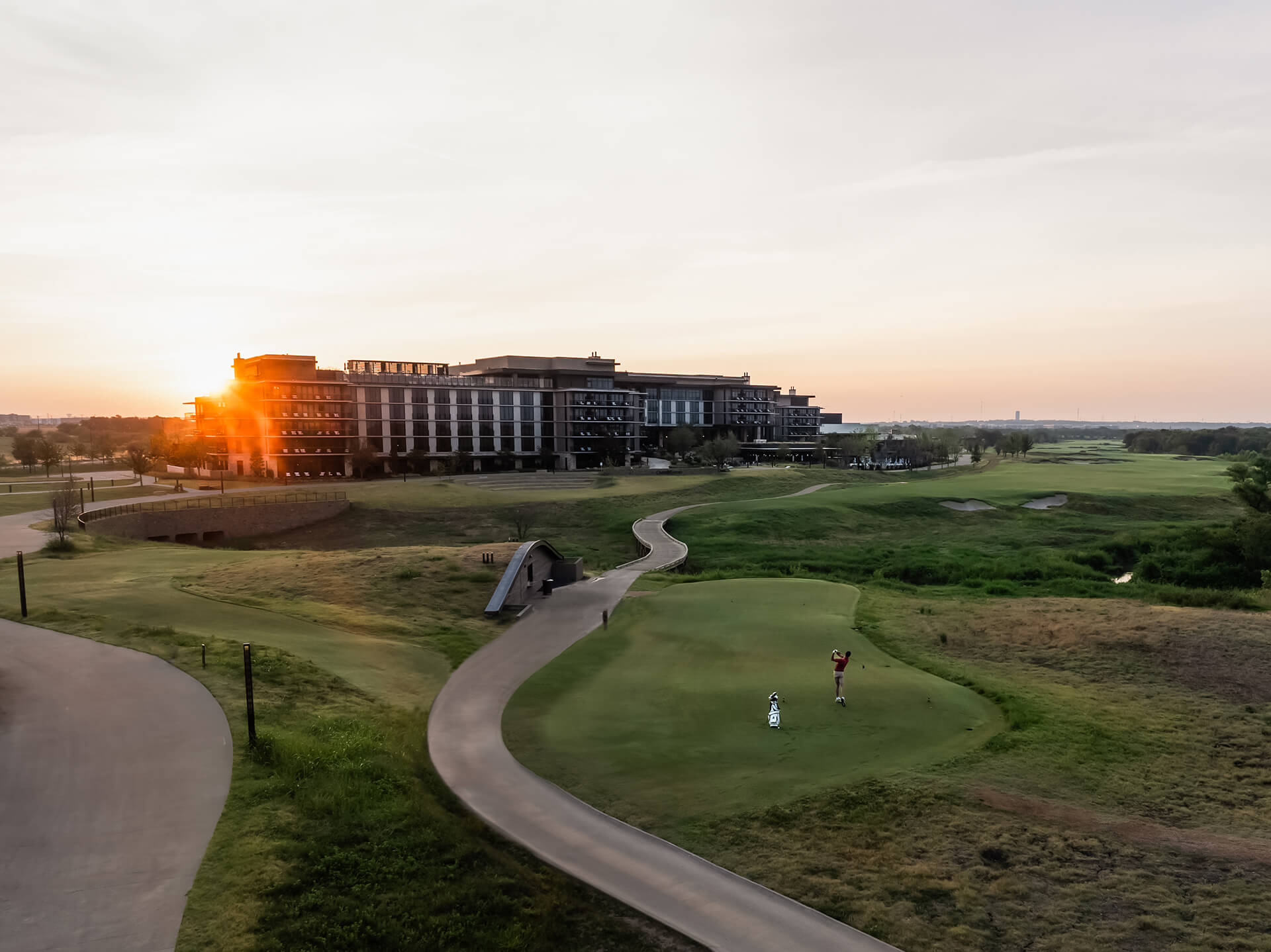 Scenic aerial view of the Omni PGA Frisco Resort hotel at sunset, overlooking a lush golf course with a golfer putting on a green in the foreground.