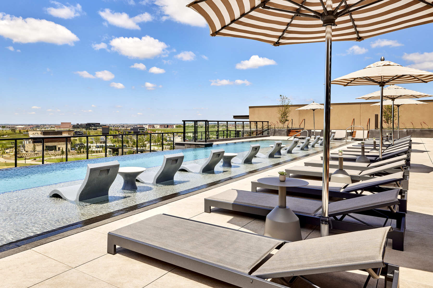 The luxurious adults-only rooftop pool at Omni PGA Frisco, featuring lounge chairs partially submerged in the water and striped umbrellas with a view of the PGA District.