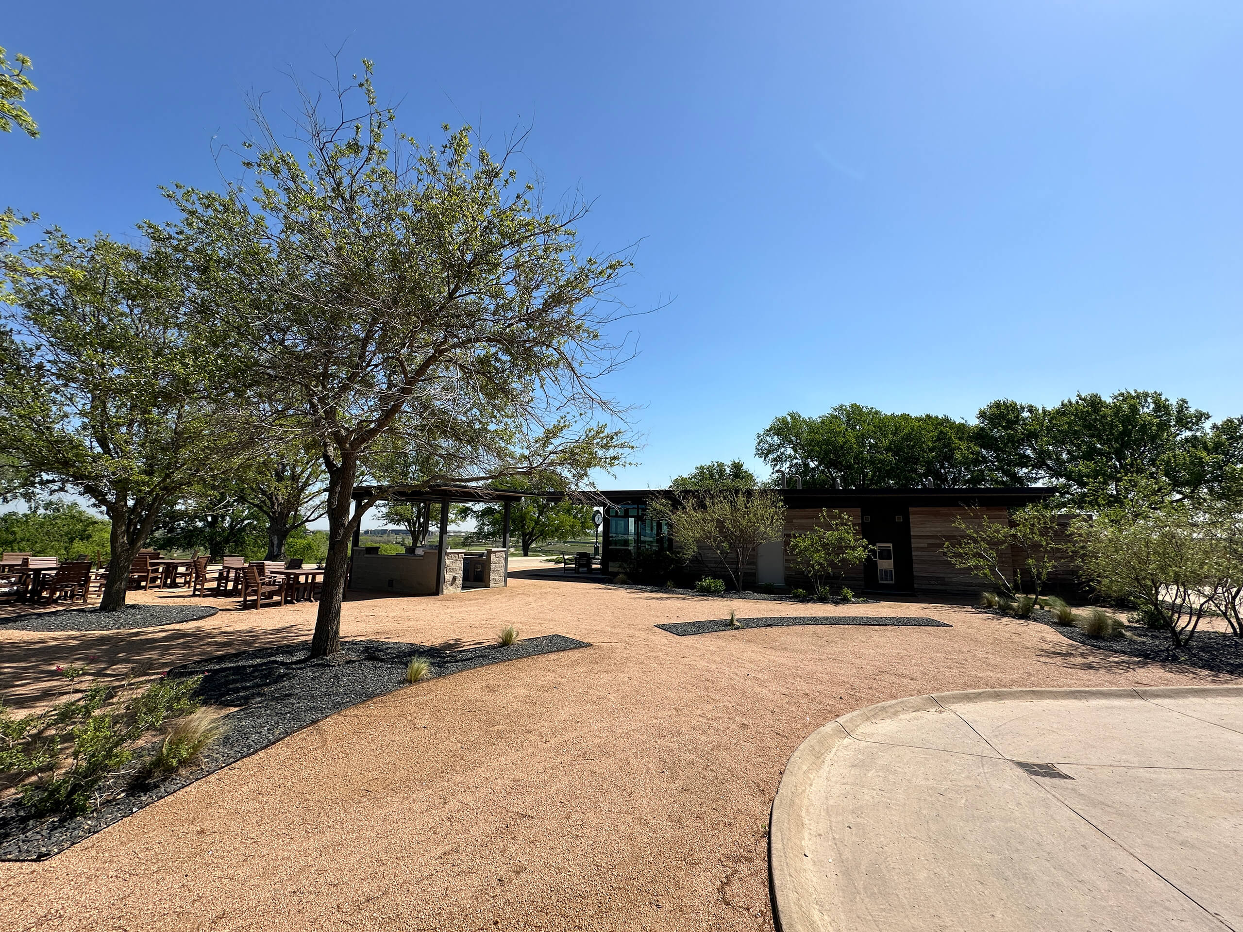 Exterior view of a modern comfort station with wood siding situated among trees and gravel paths on a golf course under a clear blue sky.