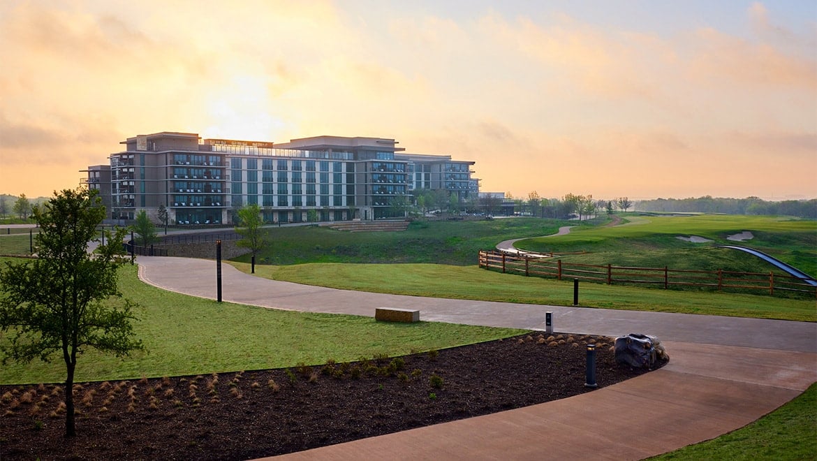 The exterior of the Omni PGA Frisco hotel, a large modern building, set on a green hill next to the golf course at sunrise.