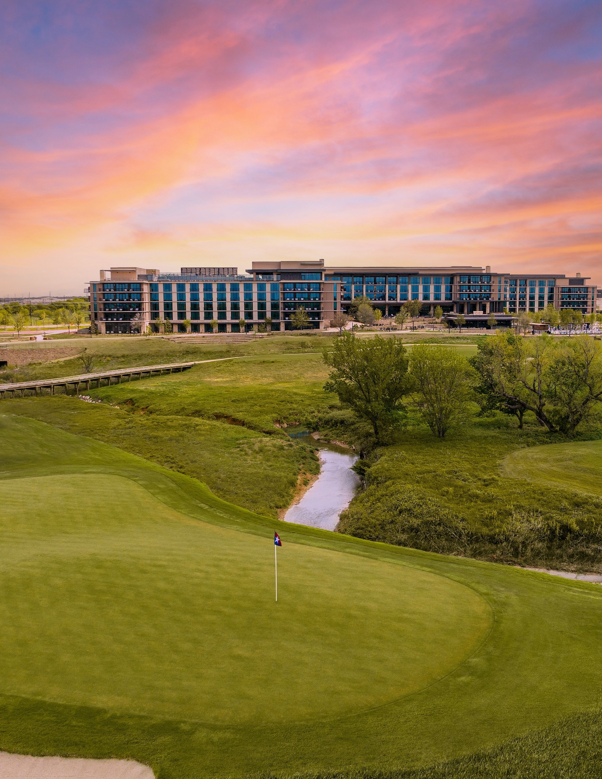 Aerial view of a golf green and a winding creek. The large Omni hotel building is visible in the distance under a pink and orange sky.