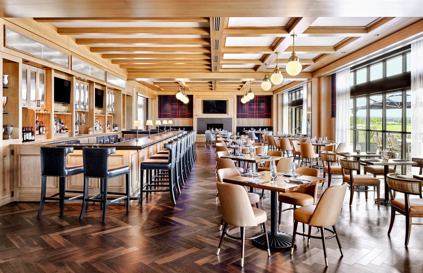 A bright restaurant with wood-paneled walls, exposed ceiling beams, a long bar, and dining tables overlooking the golf course.