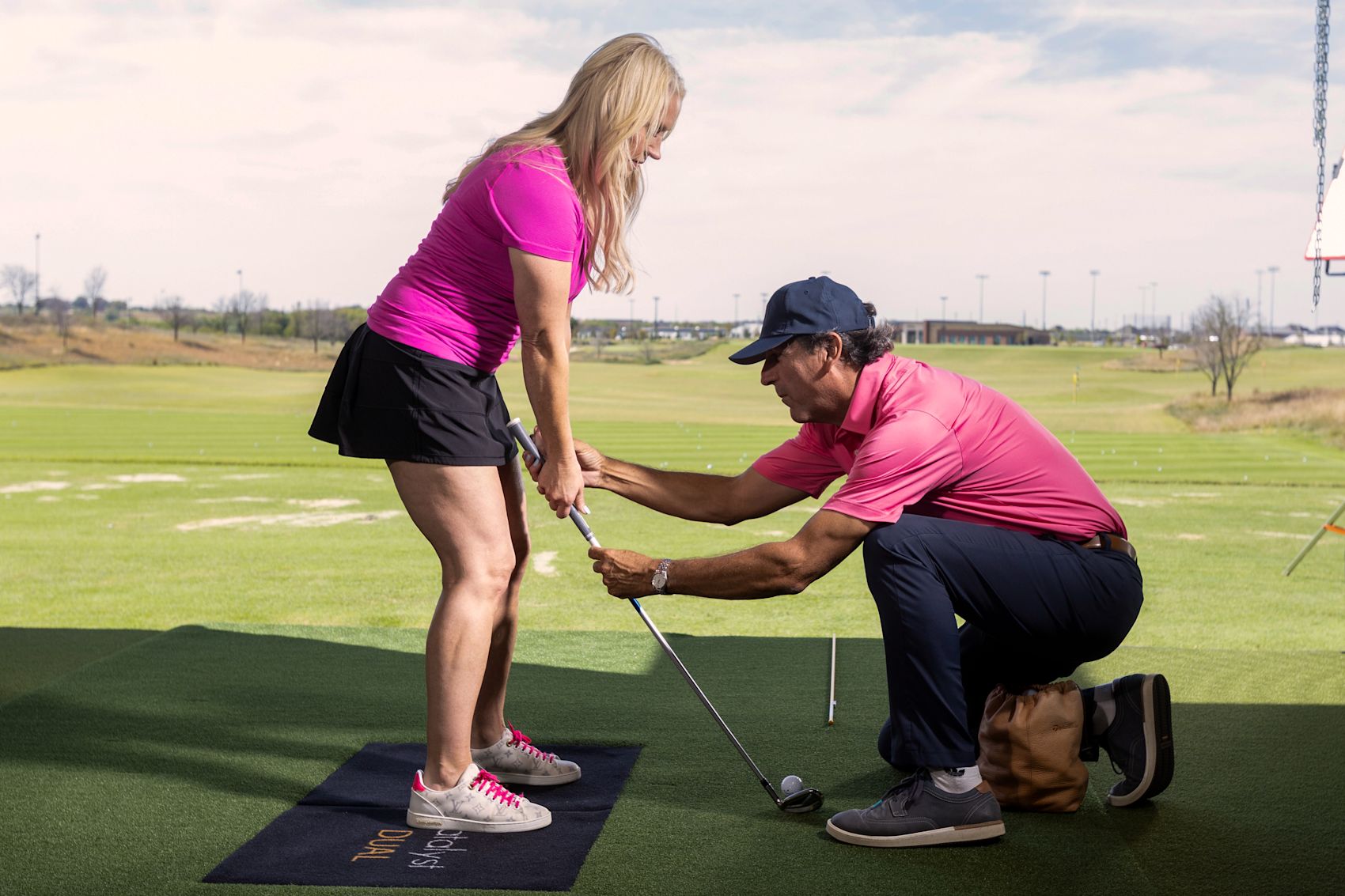 A golf coach kneels to correct a female student's grip and stance during a lesson at an outdoor driving range.