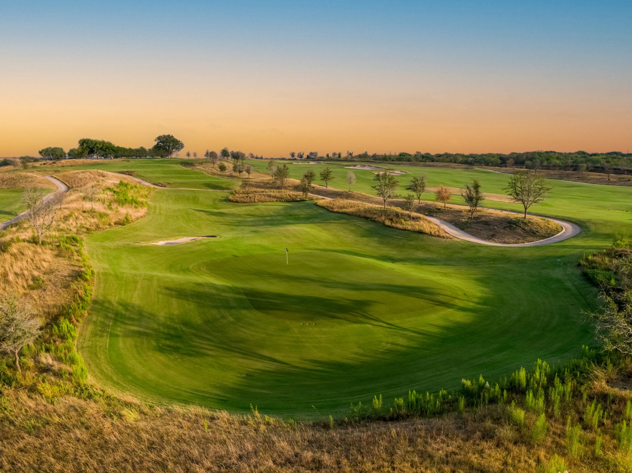An aerial view of a wide, curving golf green and fairway surrounded by natural grasses and trees at sunset.