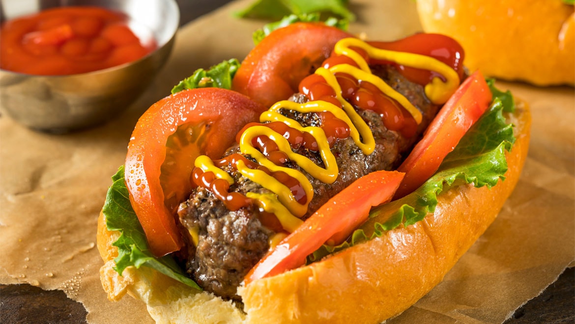 Close-up of a loaded hot dog/sausage in a bun with lettuce, tomatoes, and zigzag lines of ketchup and yellow mustard.