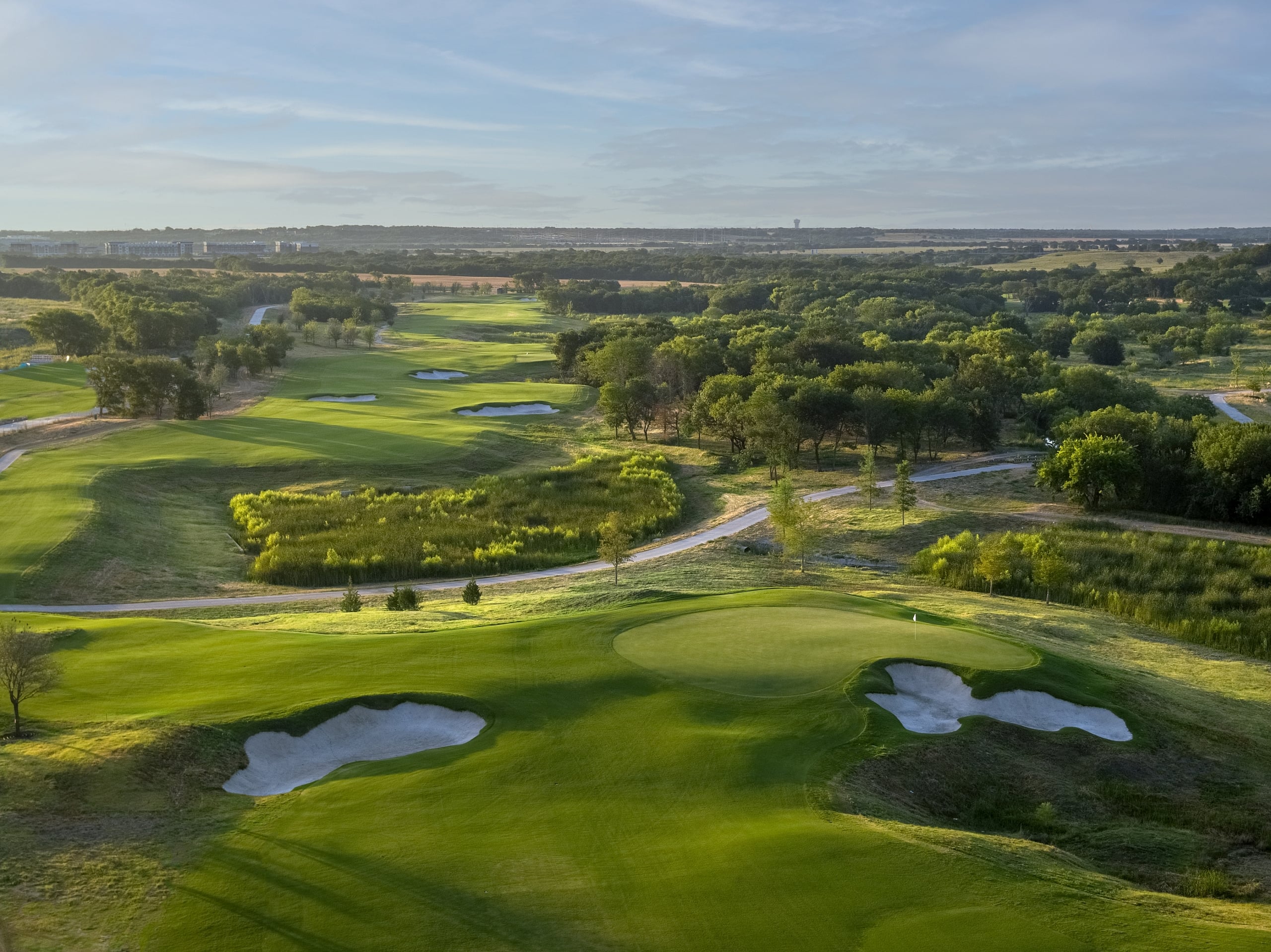 Aerial view of a sunlit, vibrant green golf course with fairways, sand bunkers, and winding cart paths through trees.