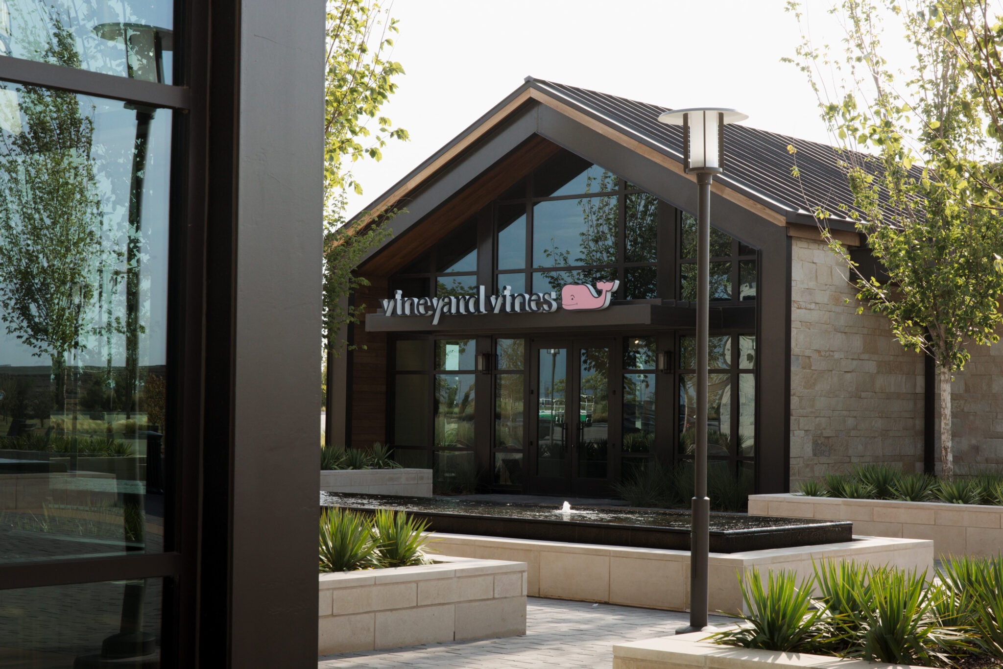 The modern exterior of a Vineyard Vines store with a gabled roof and large windows, framed by a fountain.