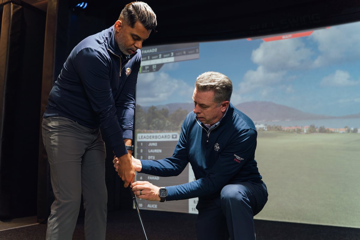 A golf coach kneels next to a student, demonstrating the correct grip on a golf club in a golf simulator bay.