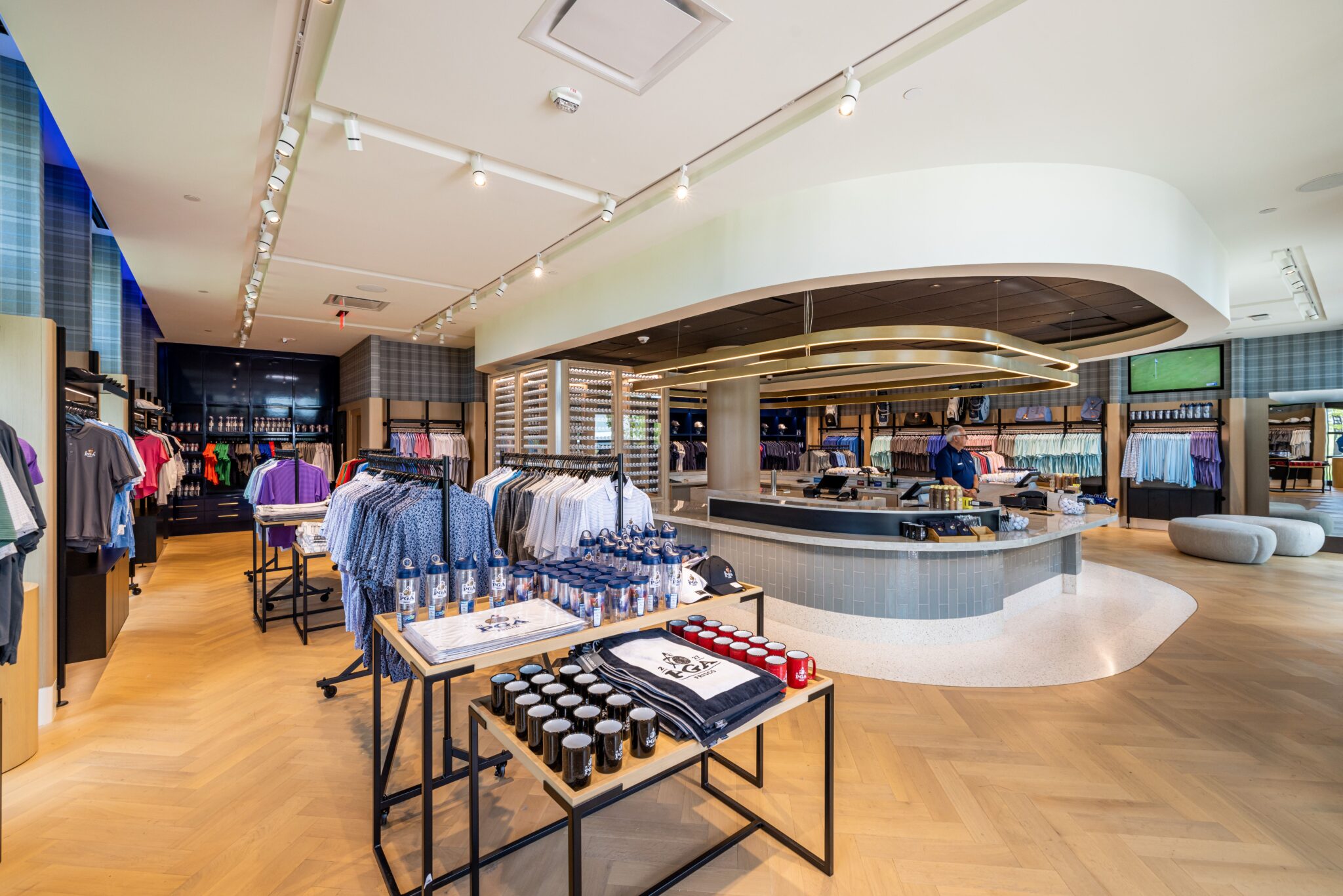 Wide view of a modern, bright golf pro shop with herringbone wood floors, apparel displays, and a curved checkout counter.