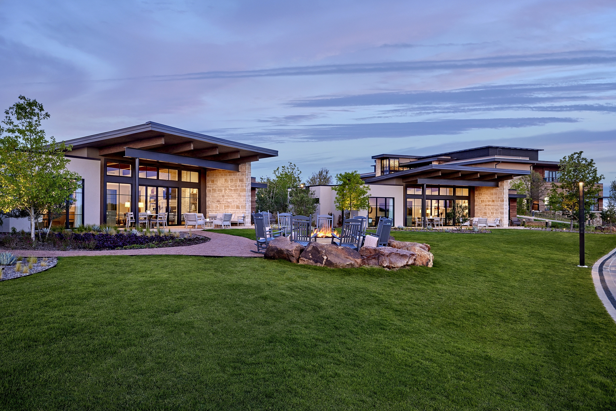 Two modern resort buildings flank a green lawn with a large rock-ringed fire pit and rocking chairs at dusk.
