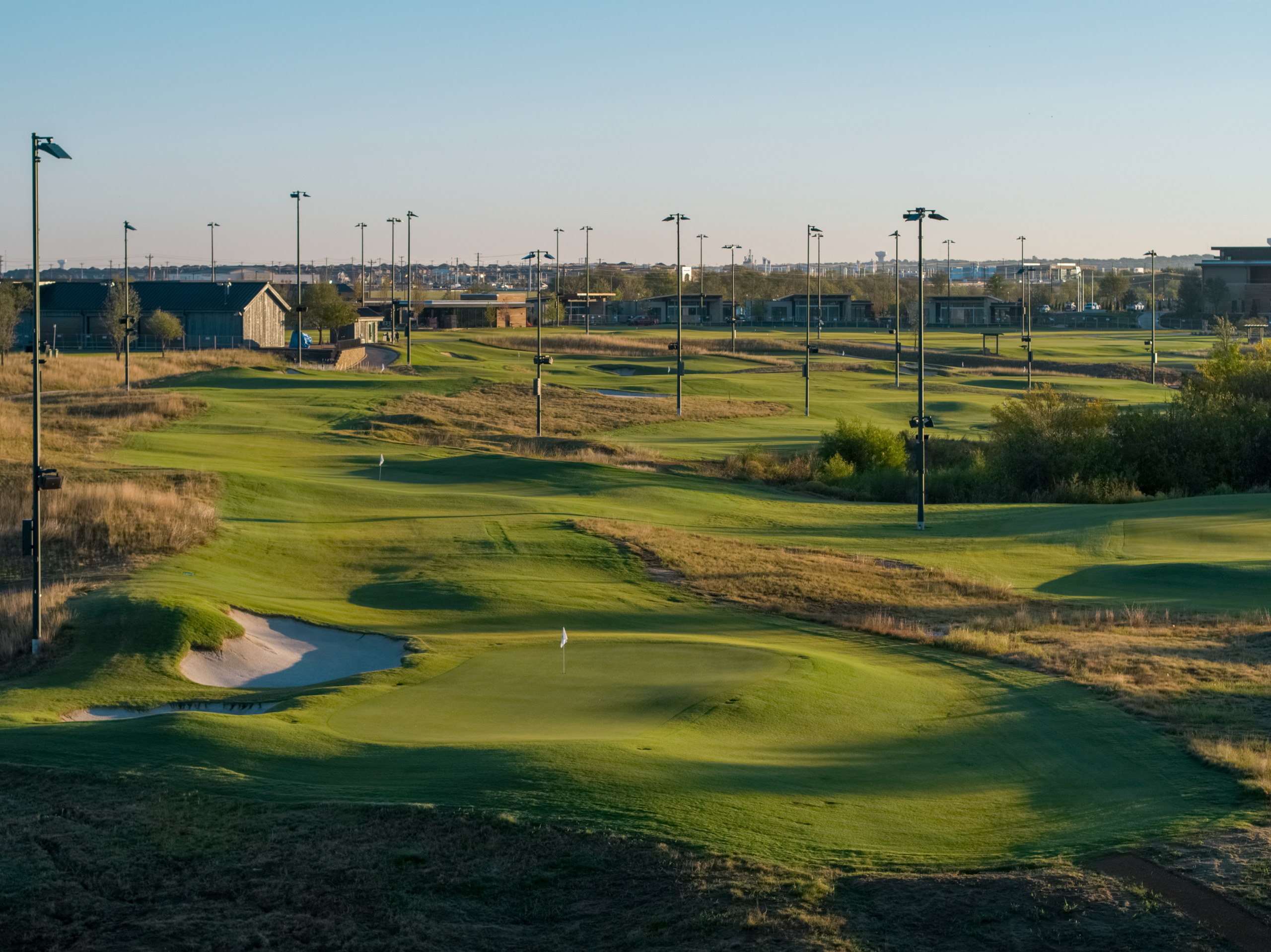 Wide view of the illuminated golf driving range and practice facility with sand bunkers and many tall light poles.