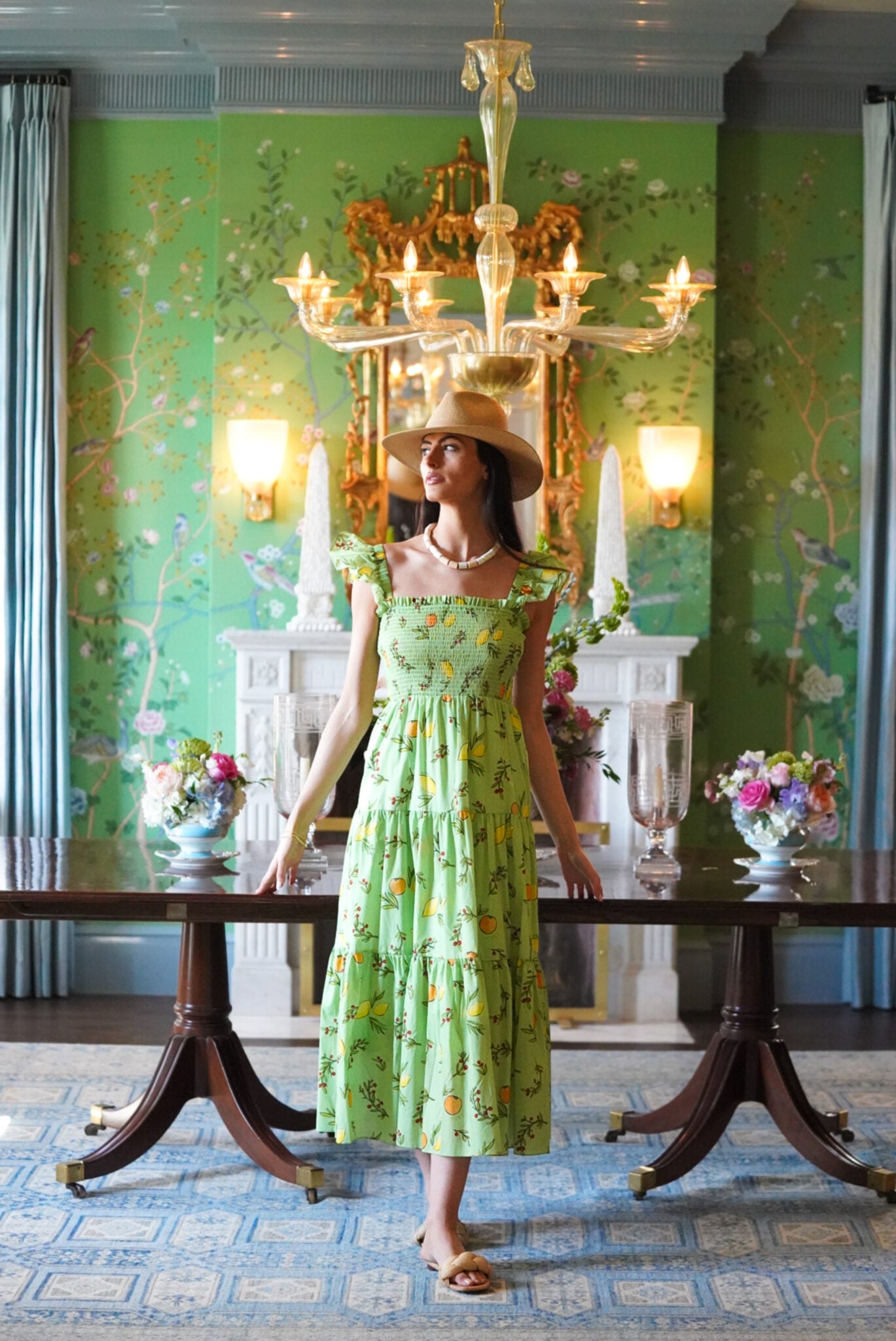 A woman in a green fruit-patterned dress stands in a room with ornate green chinoiserie wallpaper and a crystal chandelier.