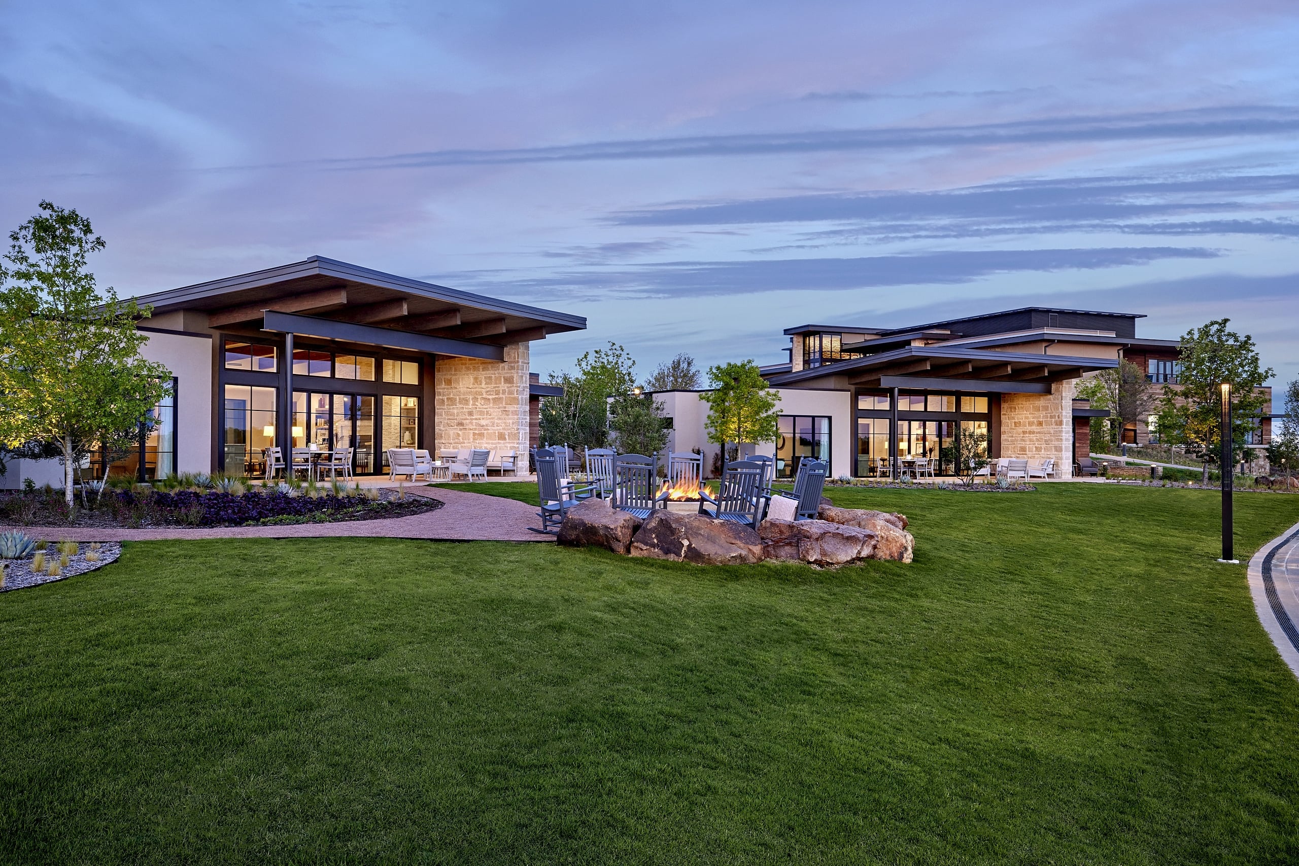 Two modern stone and glass resort buildings flank a green lawn with a large rock-ringed fire pit and rocking chairs at dusk.