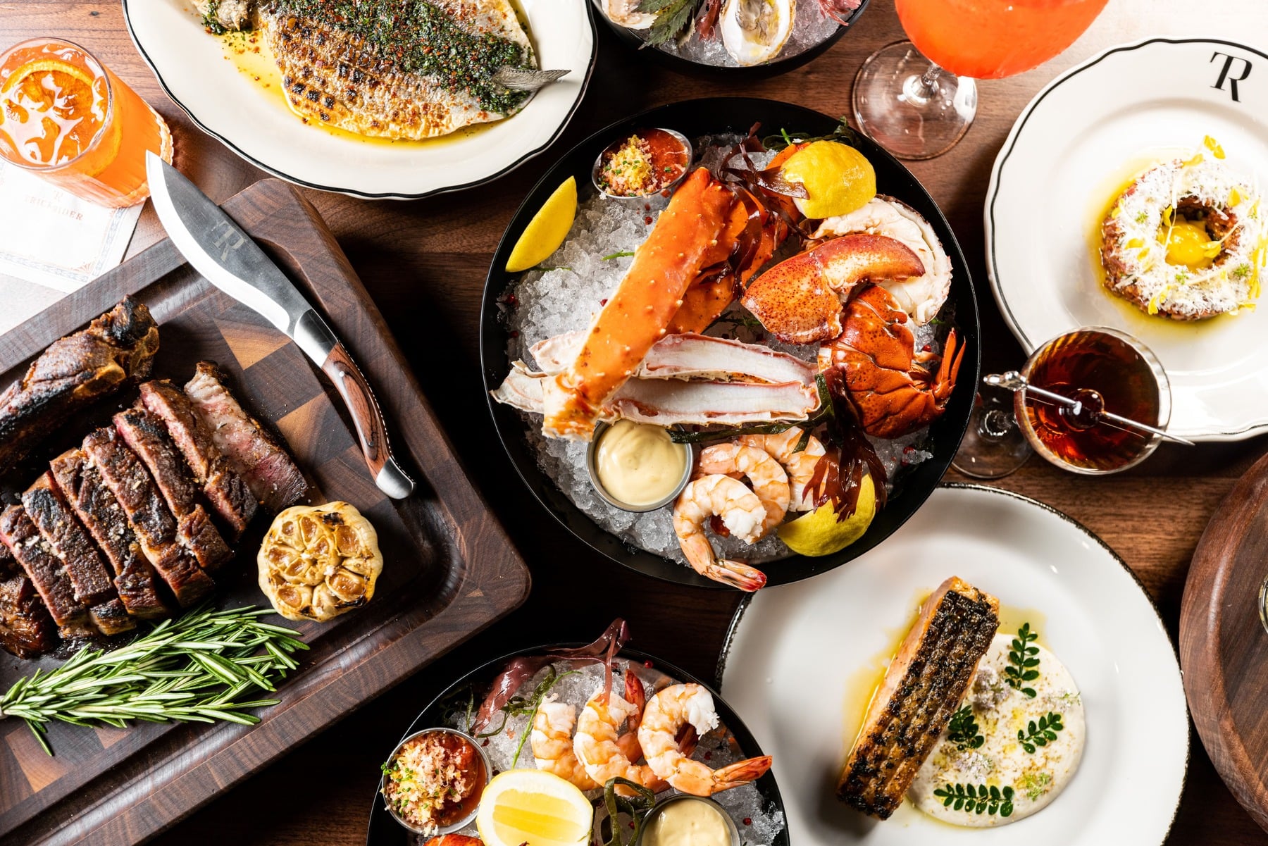 Overhead view of a restaurant spread featuring carved steak, grilled fish, chilled lobster and crab, and cocktails on a dark wooden table at Trick Rider.