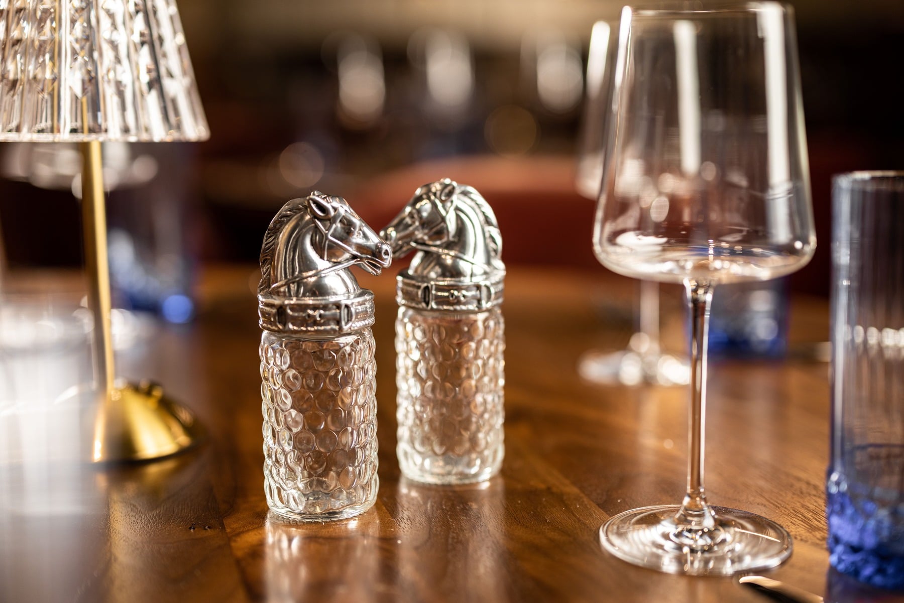 Two elegant glass salt shakers with silver horse-head tops, set on a dark wood table next to wine glasses in the Trick Rider restaurant.