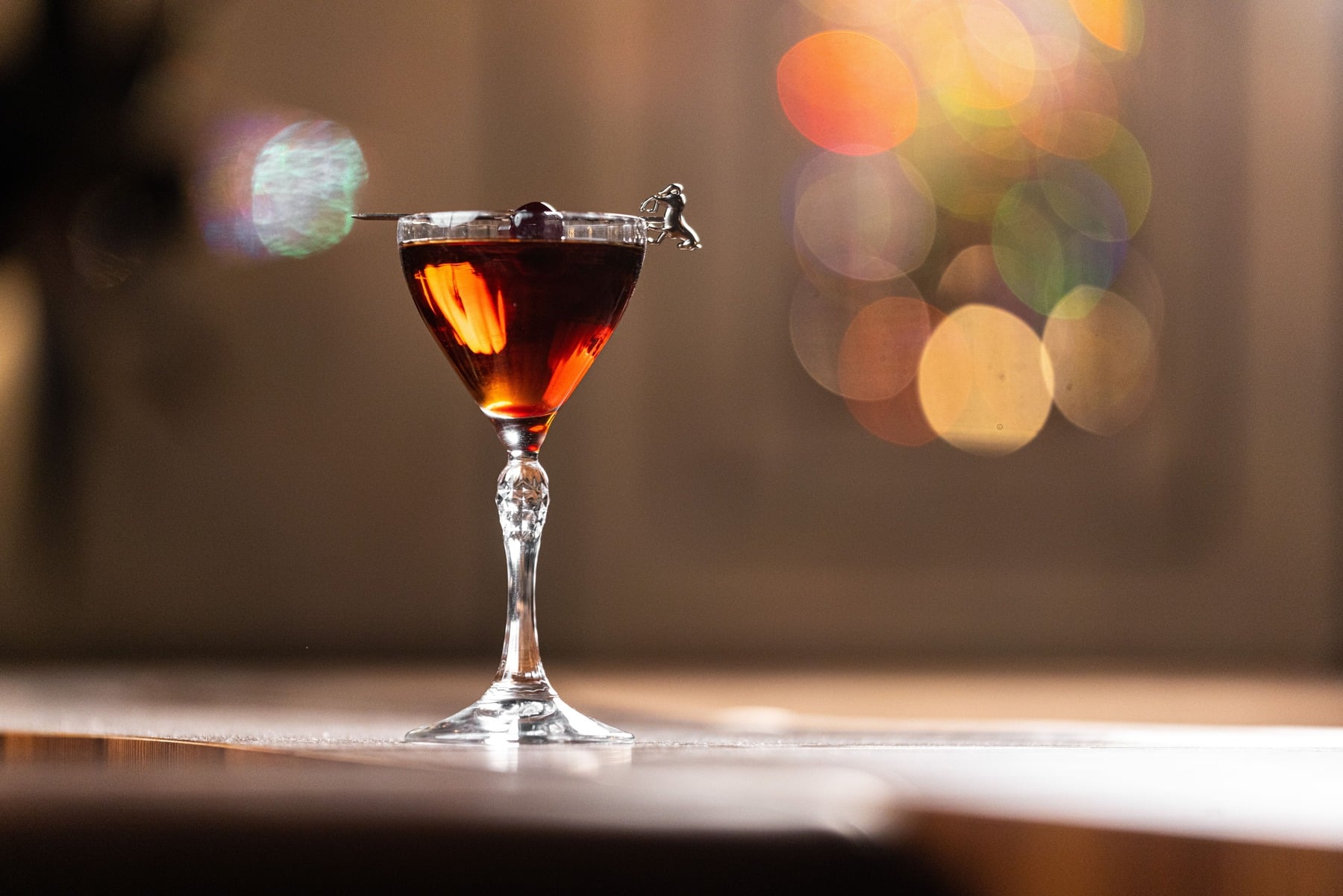A dark, moody photo of a cocktail in a coupe glass, garnished with a cherry and a small metal golf-themed stirrer, with blurred bokeh lights in the background.