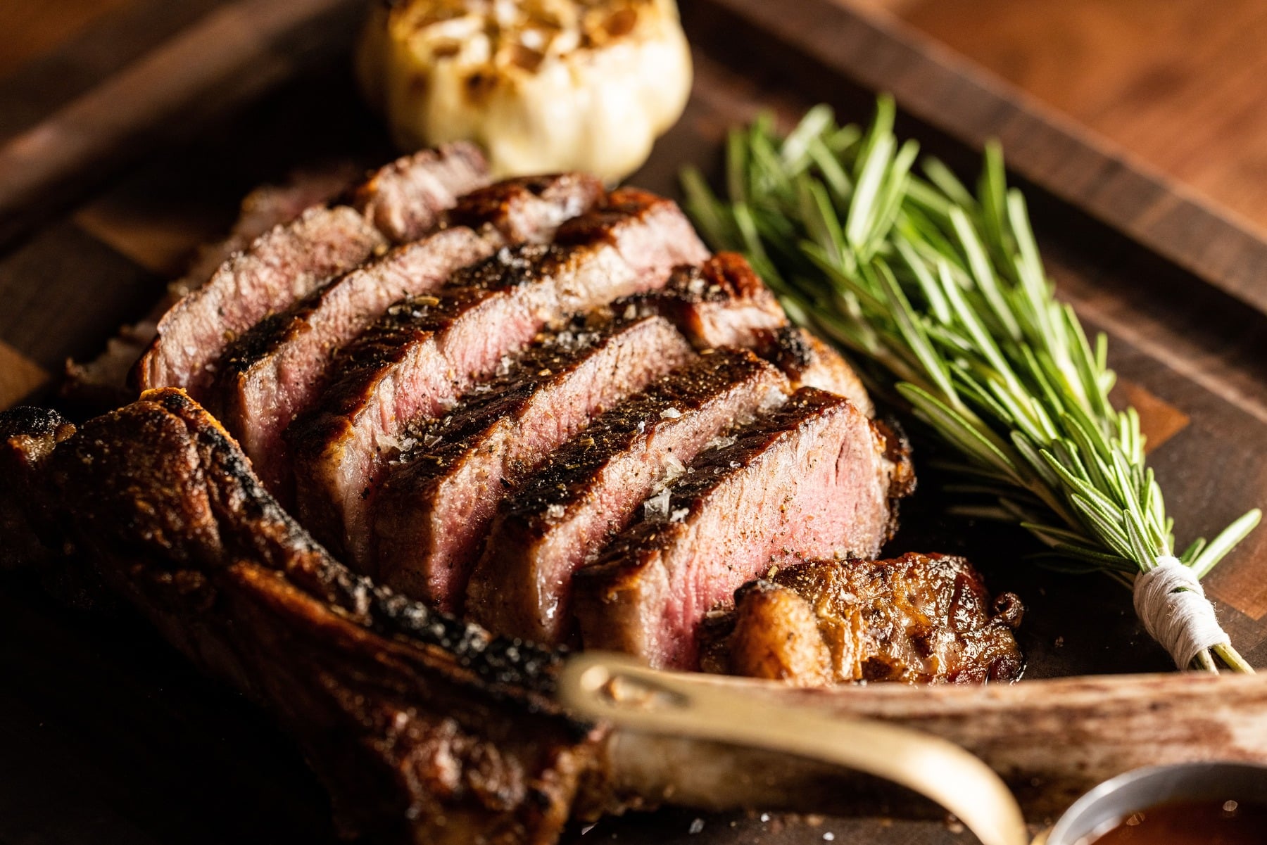 A close-up of a carved ribeye steak served on a wooden board, garnished with a bundle of fresh rosemary and roasted garlic.