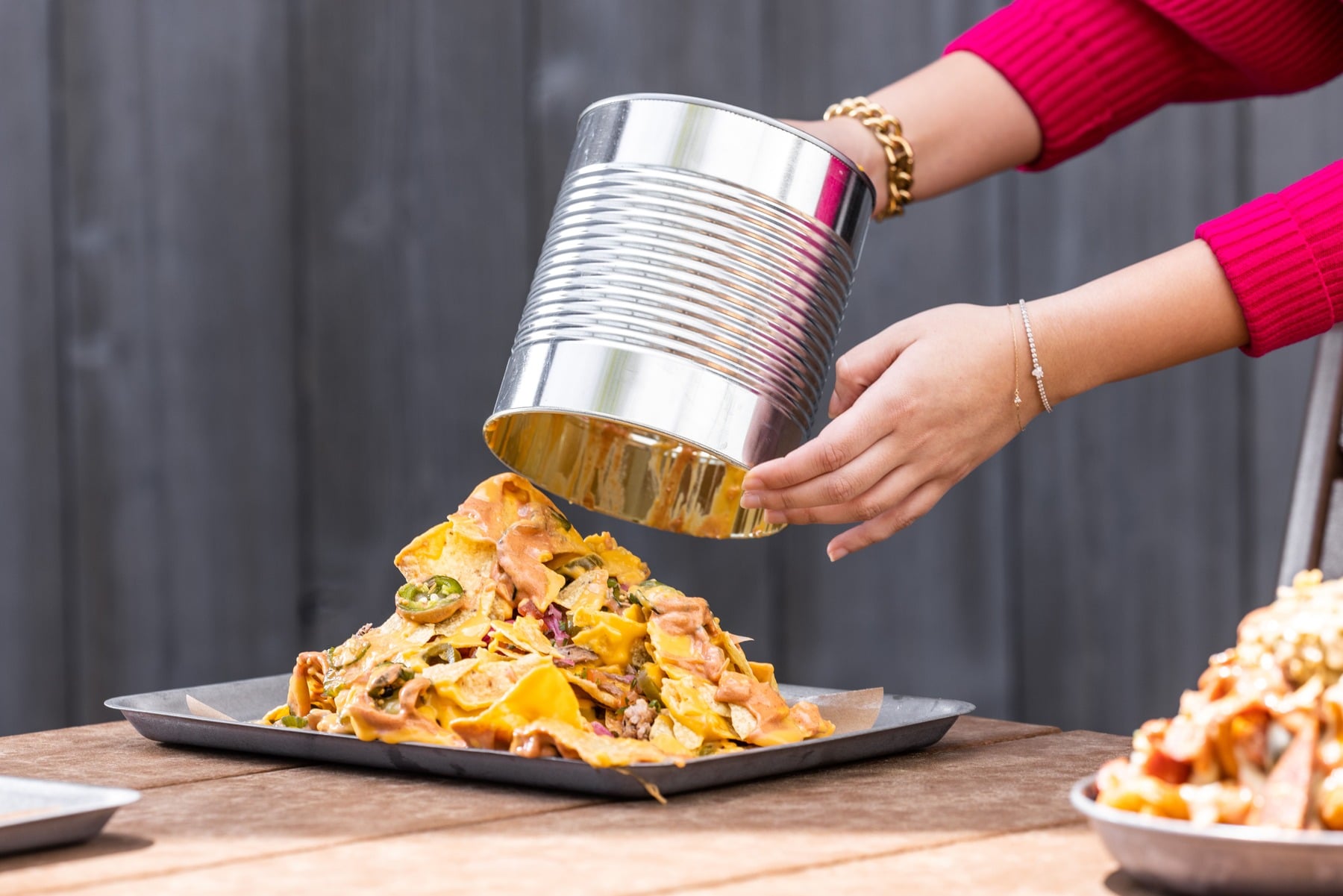 Hands pouring the contents of a large metal can over a pile of nachos or layered tortilla chips served on a baking tray.
