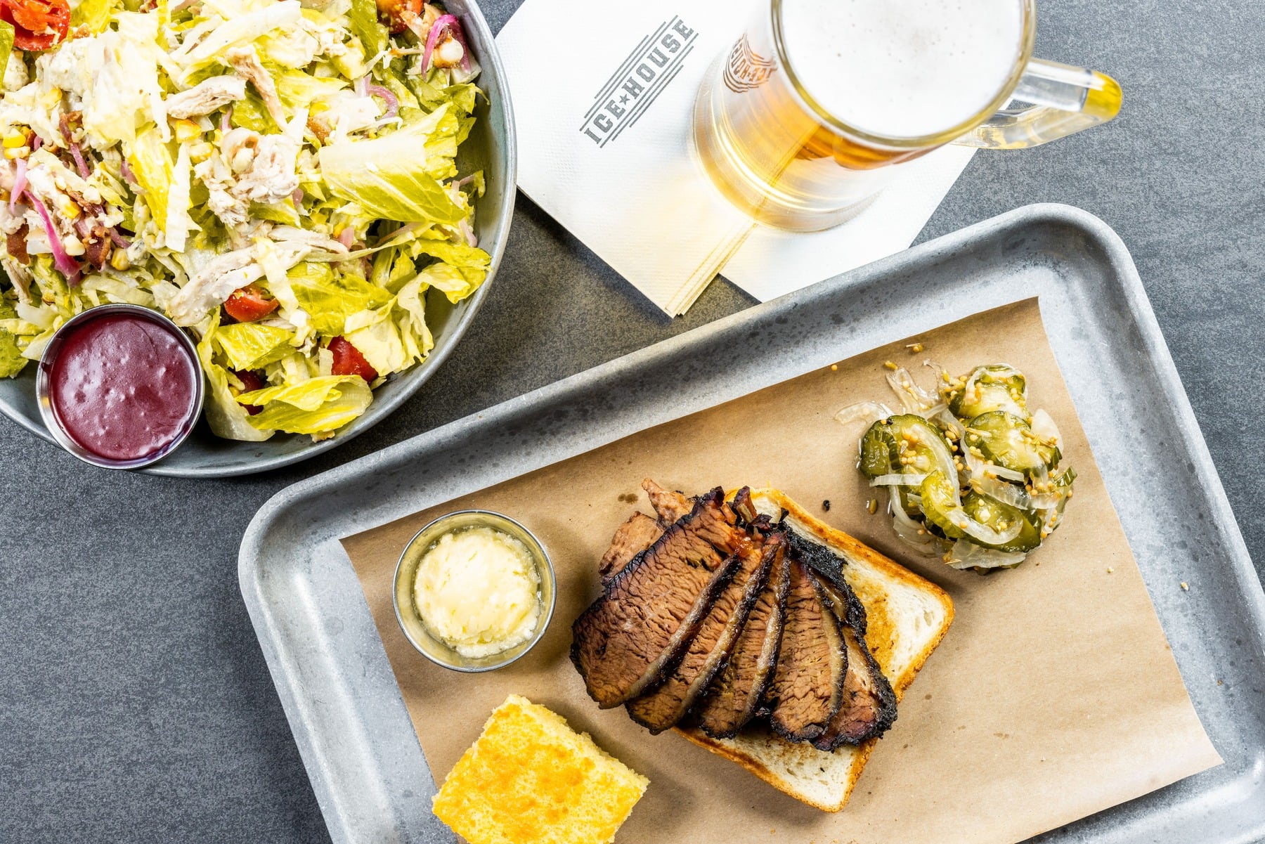 Overhead view of a tray of sliced smoked brisket and cornbread, next to a bowl of fresh salad and a mug of beer, served at Ice House.