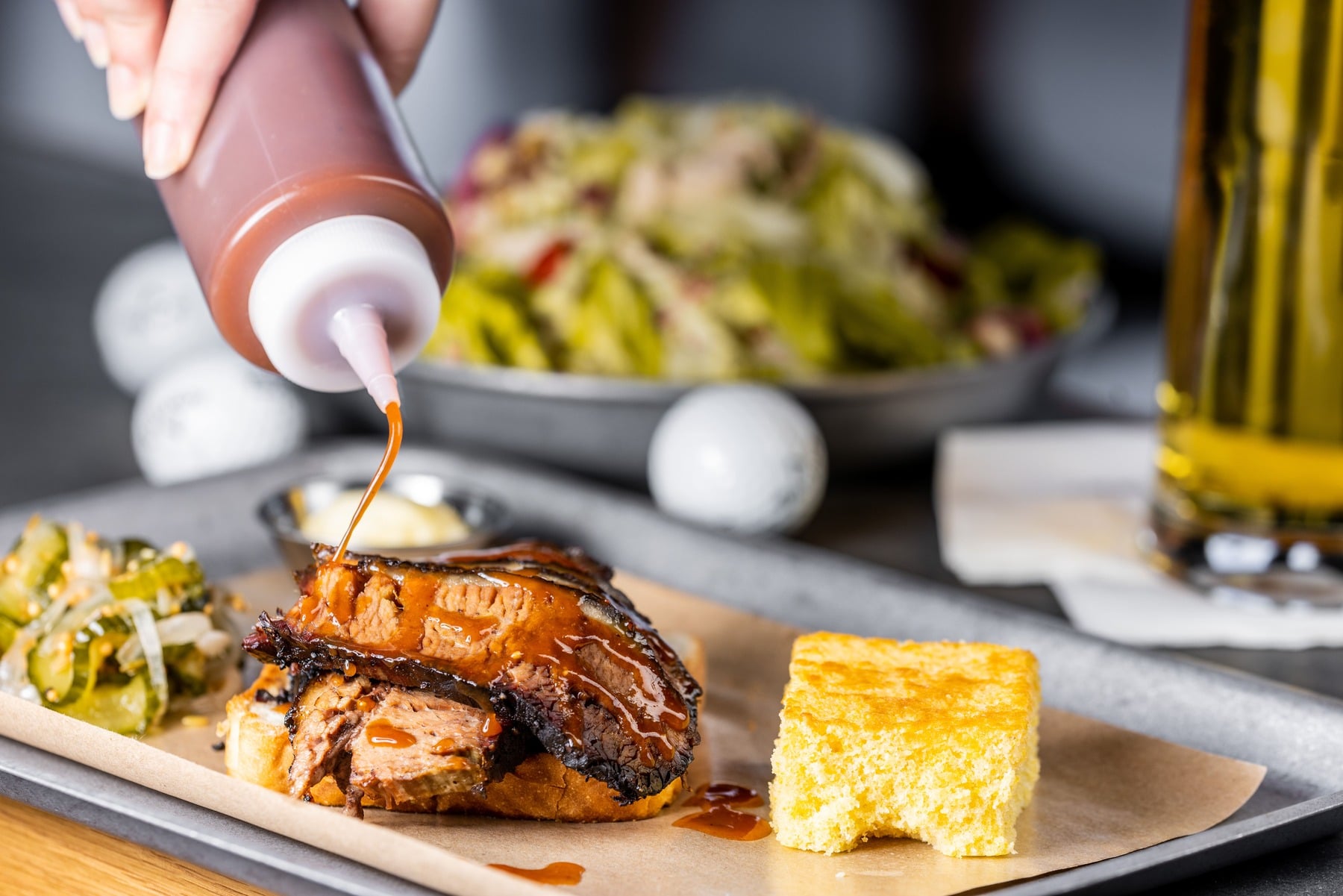 Close-up of sliced smoked brisket served on toasted bread with a side of corn bread, being drizzled with barbecue sauce.