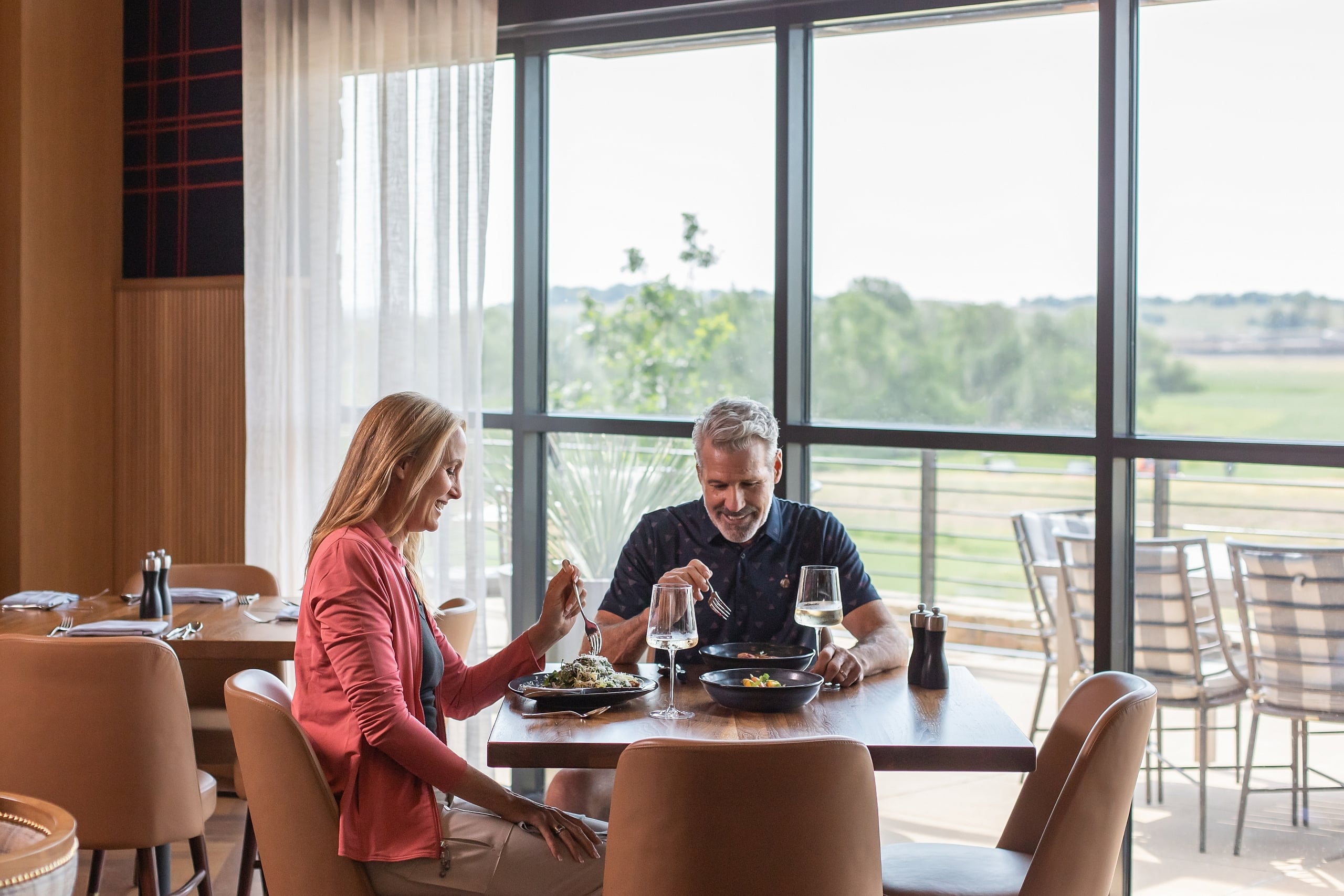 A smiling couple enjoys a meal and white wine at a dining table next to large windows overlooking a green landscape.