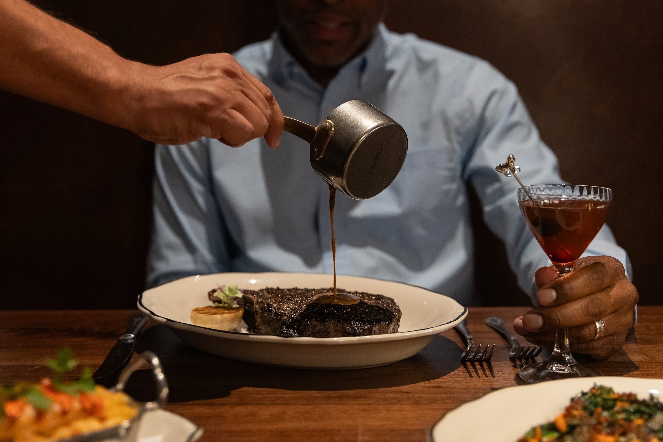 A server pours sauce over a dark steak on a plate while the diner holds a cocktail in a dimly lit restaurant.