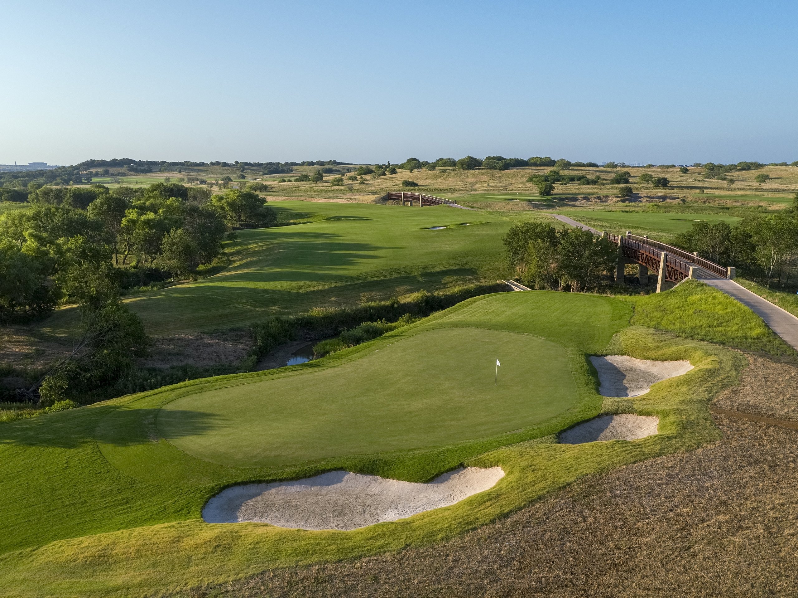 A golf green with a flag and sand bunker, showing a wide, elevated wooden bridge spanning the course in the distance.
