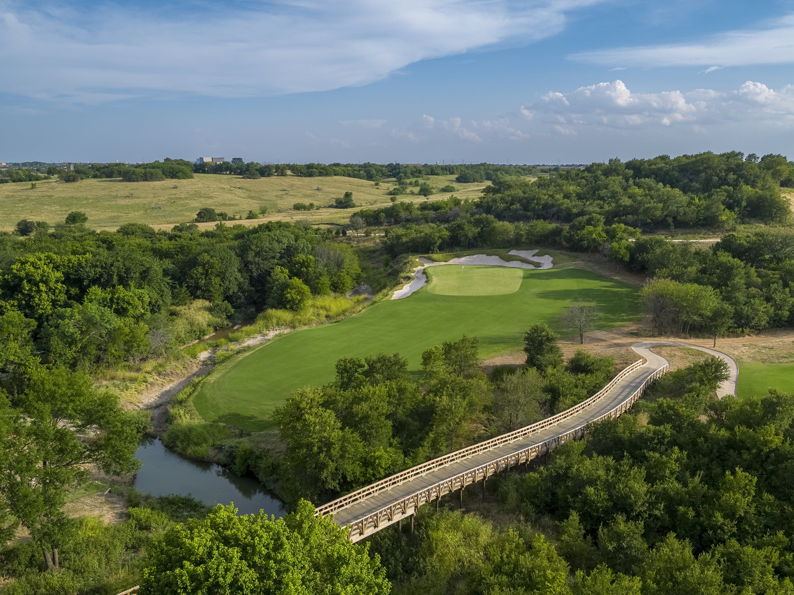 Aerial view of a green golf hole with a sand bunker, connected to a cart path by a long, wooden bridge over a creek.
