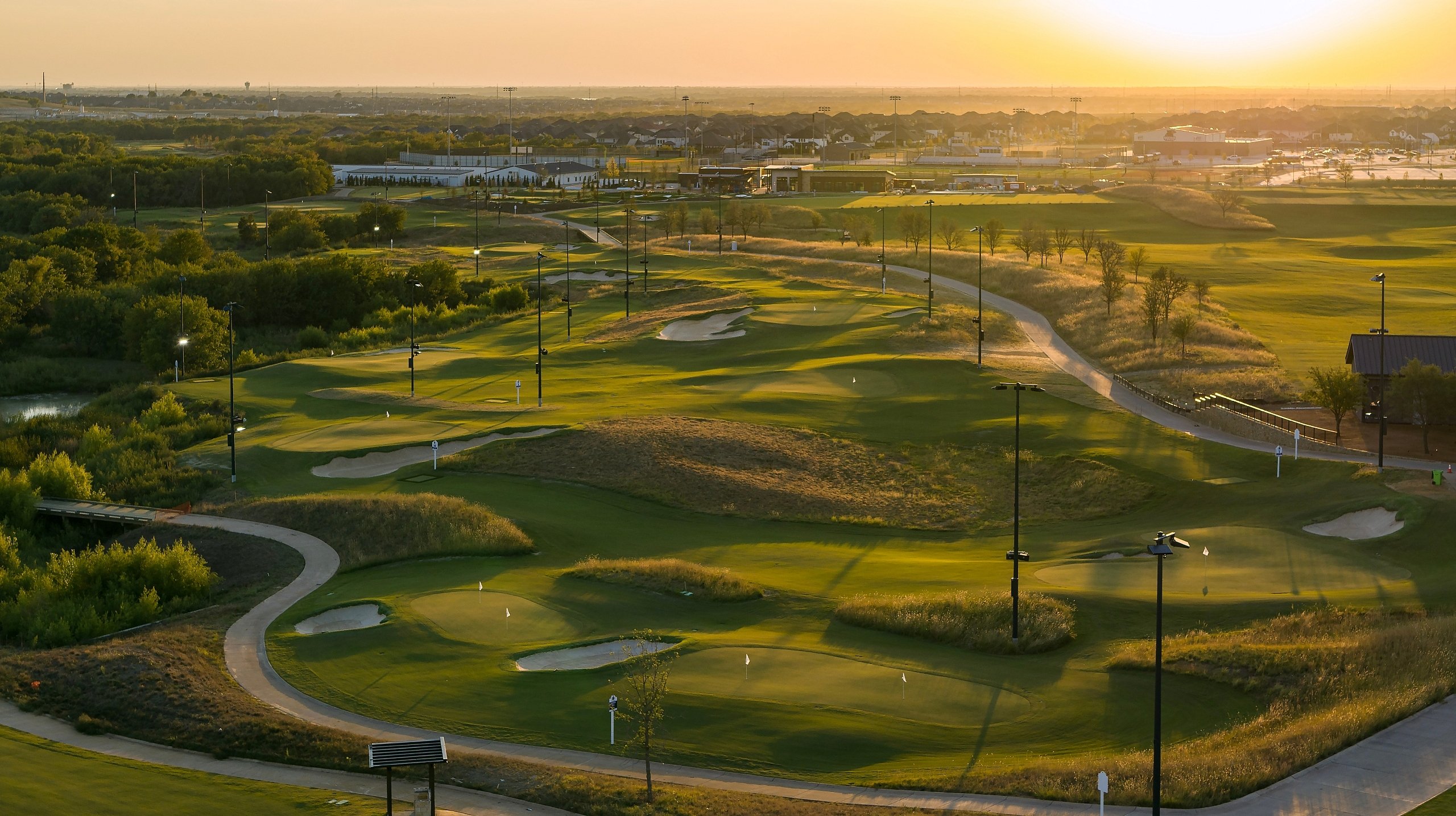 Aerial view of a sprawling golf course and driving range with bunkers and cart paths, bathed in the golden light of sunset.