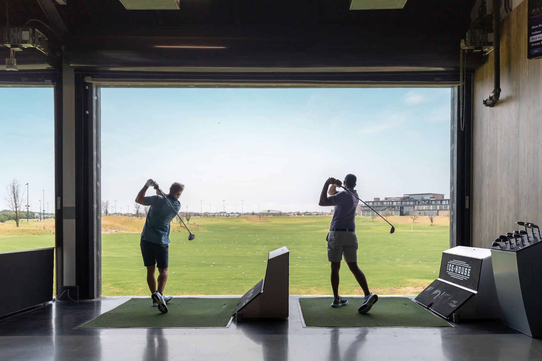 Two men practice their golf swings in an open-air driving range bay at the Ice House, with a wide view of the driving range and landscape in the background.