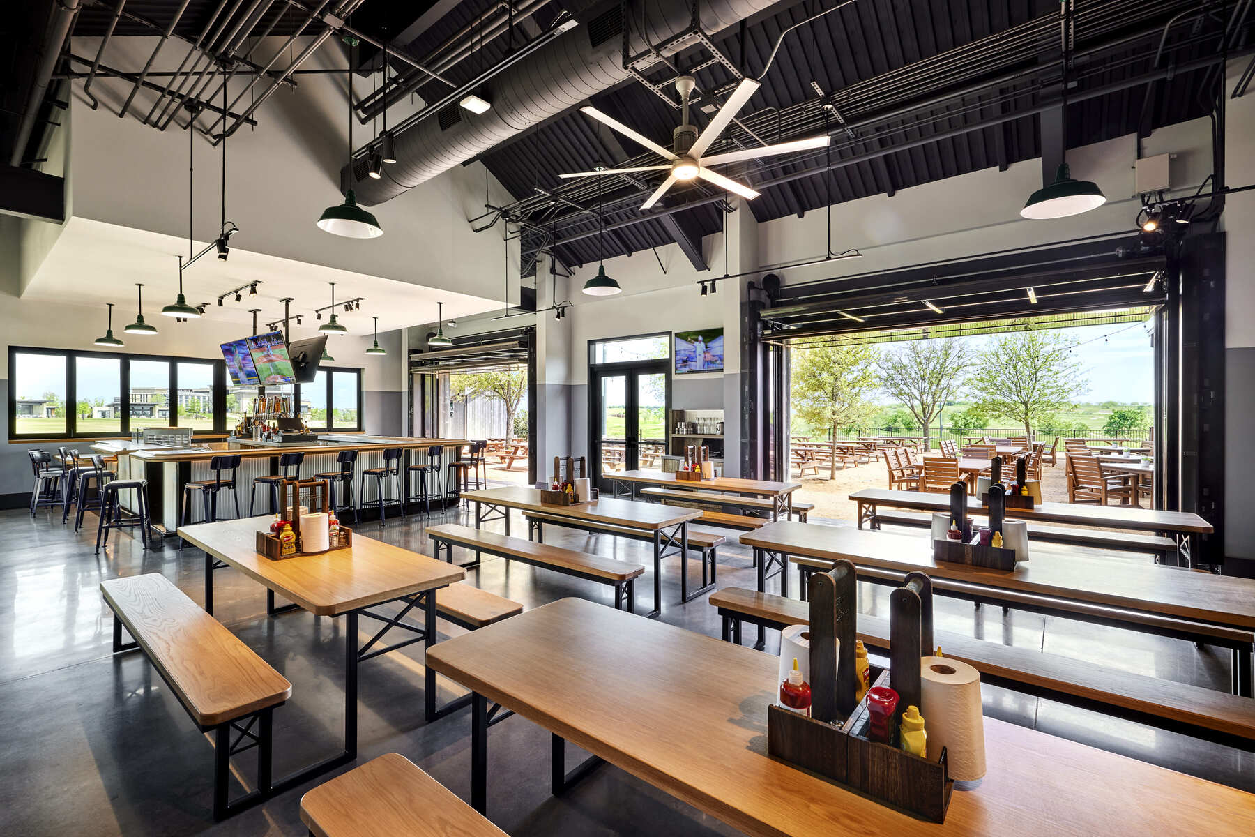 Interior view of a large, casual restaurant/bar area with high ceilings, multiple wooden picnic-style tables and benches, an open bar area, and large garage-style doors leading to an outdoor patio with picnic tables.