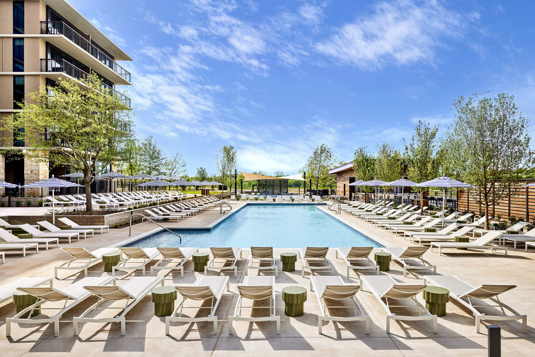 Wide view of the Omni PGA Frisco Resort Leisure Pool, showing rows of modern lounge chairs, umbrellas, and the hotel building facade.