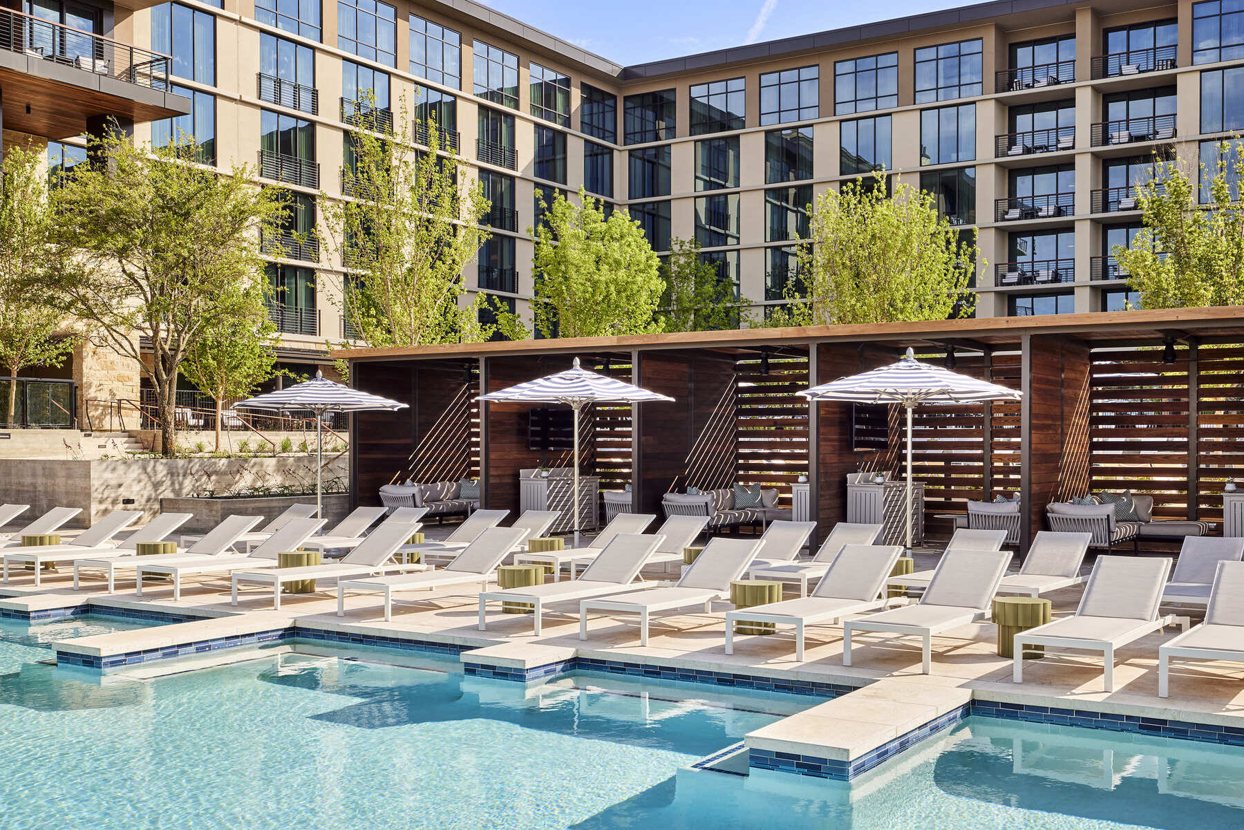 The pool deck at the Omni PGA Frisco Resort showing cabanas with slatted wood walls, lounge chairs, and striped umbrellas lining the pool edge.