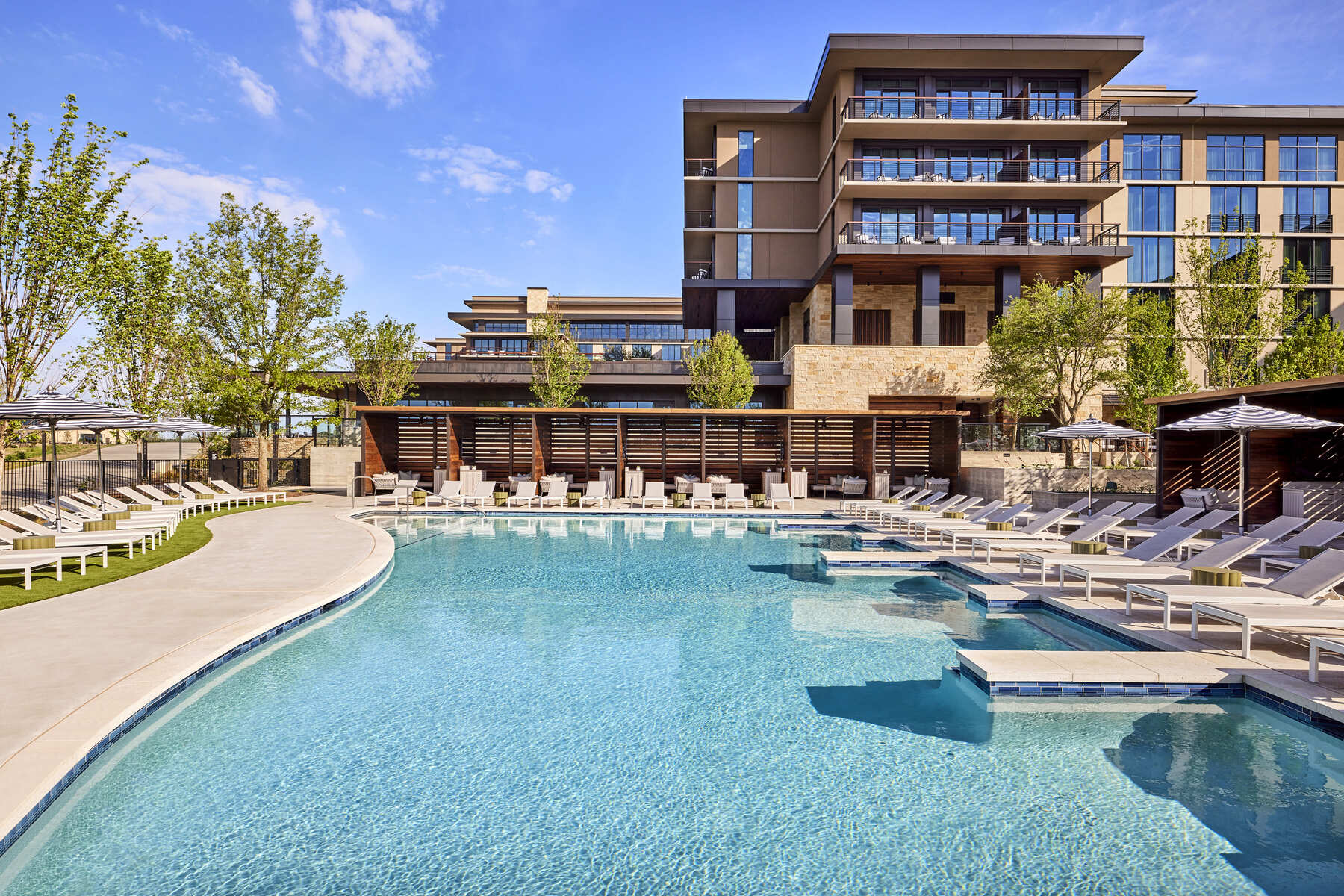 A sunny view of the outdoor leisure pool at the Omni PGA Frisco Resort, featuring lounge chairs lined up on a shallow water ledge.