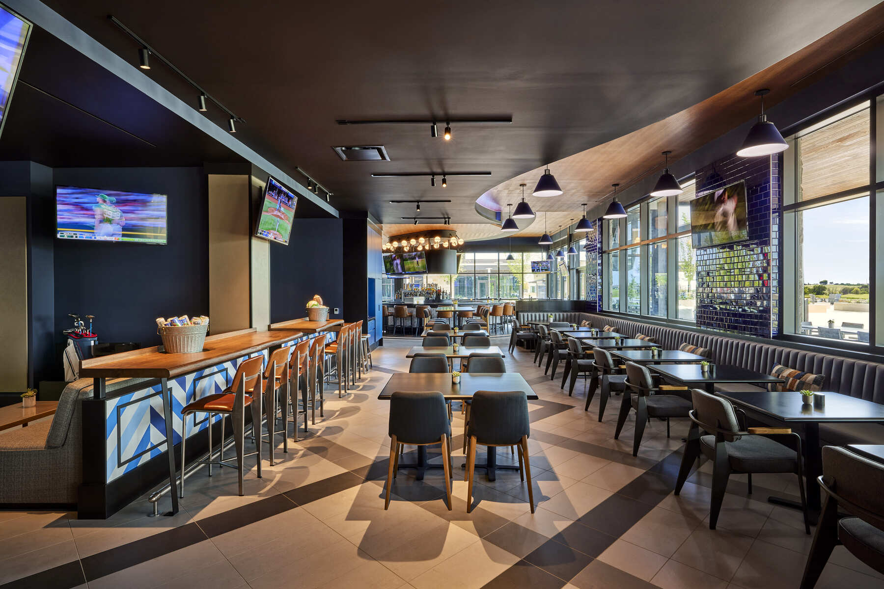 Interior view of Lounge by Topgolf at Omni PGA Frisco Resort, showing the bar, high-top seating, dining tables, and large windows with sports visible on screens.